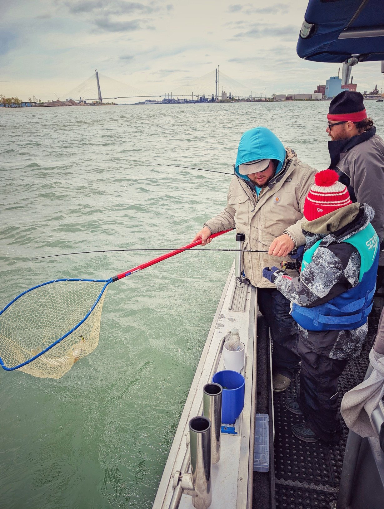  A young angler reeling in a fish while a Reel Fish'n guide readies the net on the Detroit River, with the Ambassador Bridge visible in the background. 