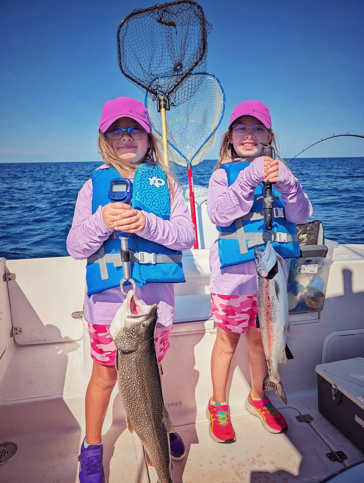  Two young girls in life jackets proudly posing with their Lake Trout catch during a family-friendly Oscoda fishing charter on Lake Huron. 