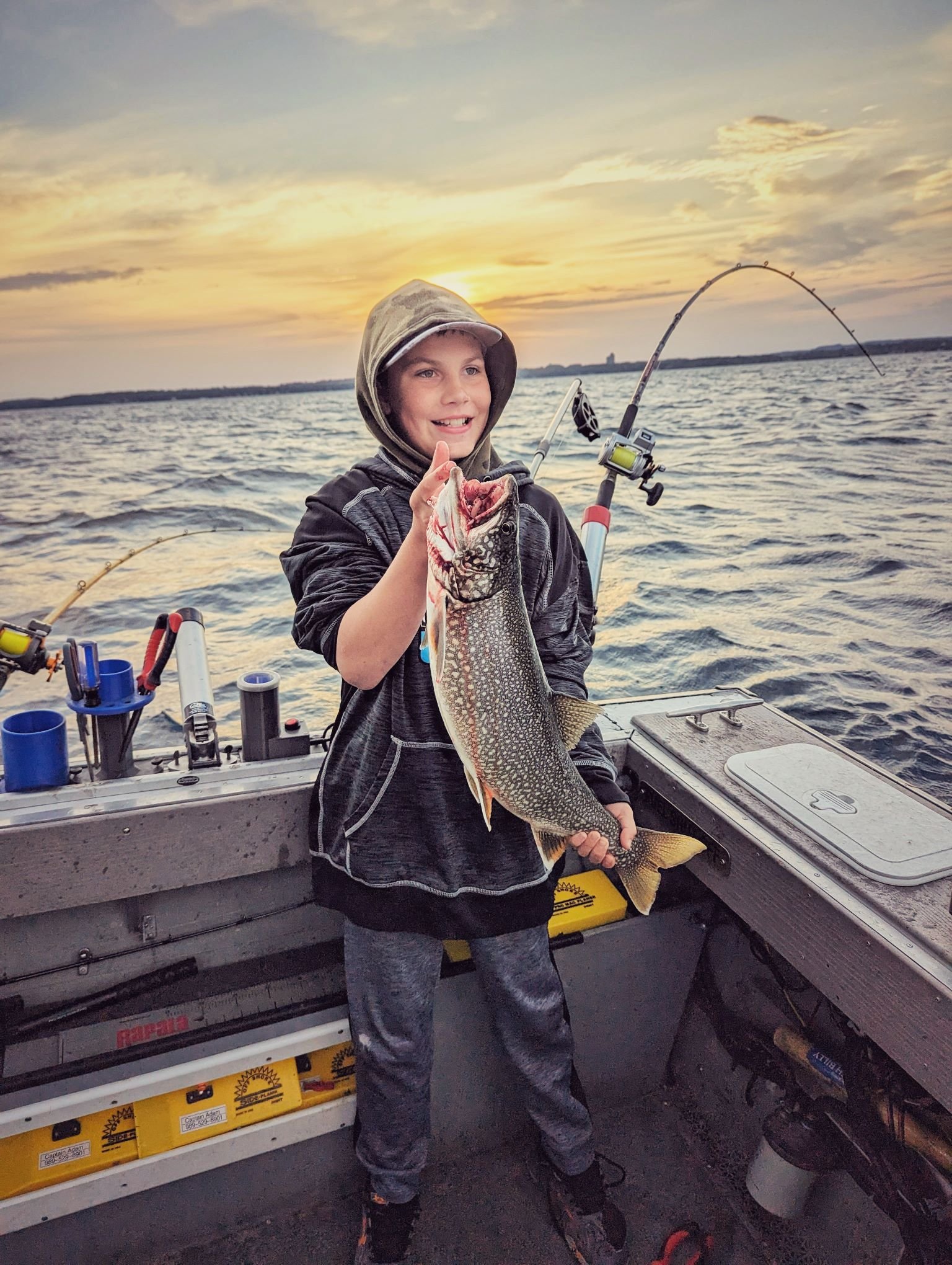 A young angler holding a fresh Lake Trout with a second rod doubled over in the background during a high-action sunrise charter on Grand Traverse Bay.   