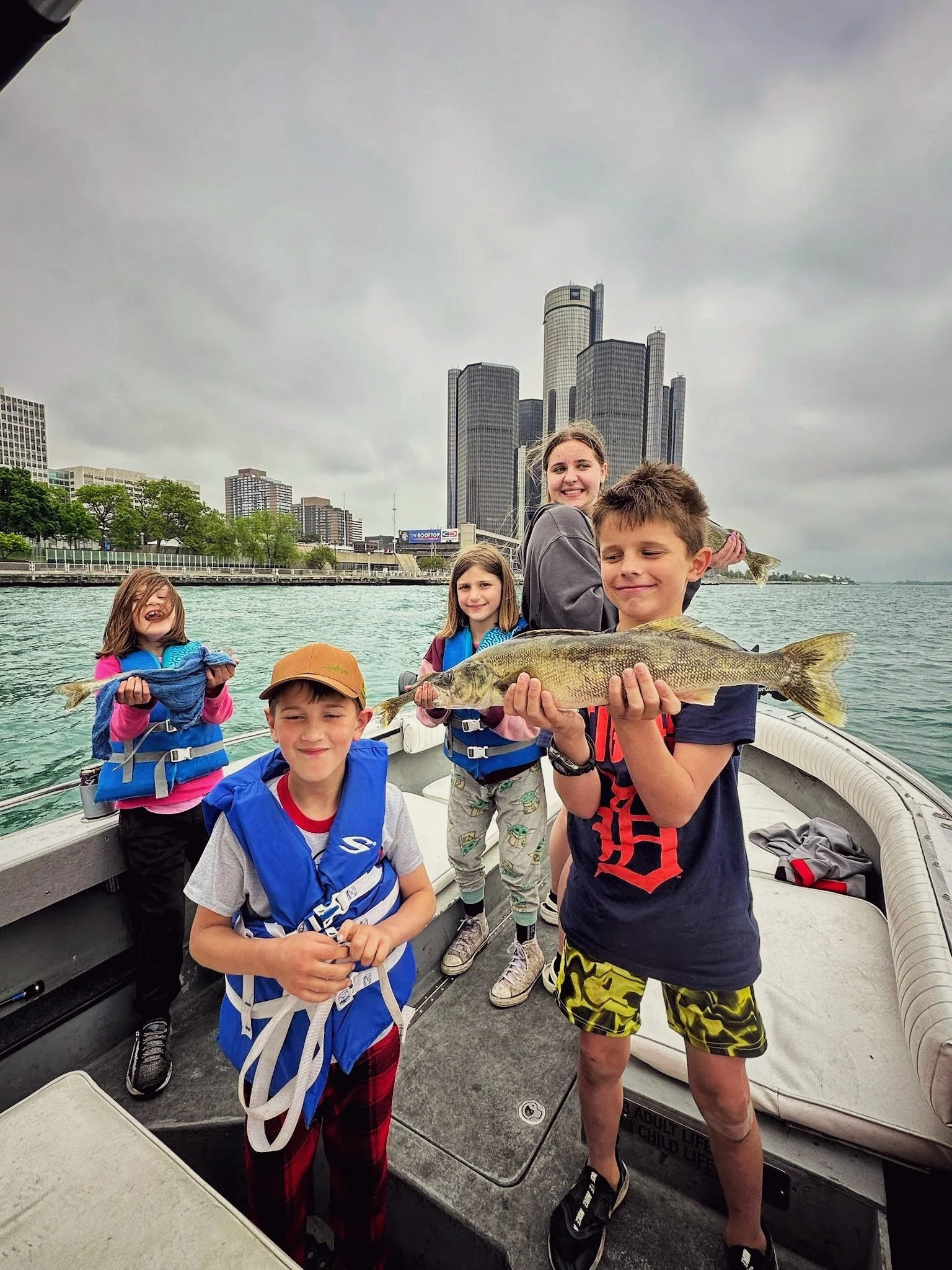  A group of children proudly posing with a large walleye on a Reel Fish'n charter boat with the Detroit Renaissance Center and city skyline in the background. 
