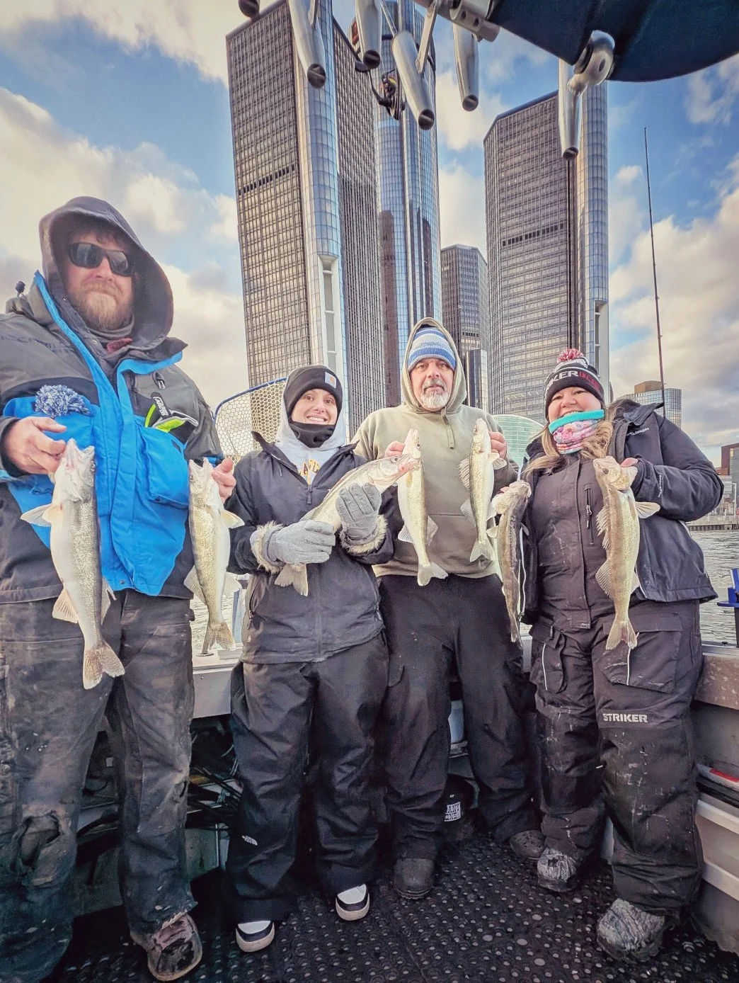  A group of four anglers in winter fishing gear holding up fresh walleye on a Reel Fish'n charter boat with the Detroit skyline towering in the background. 