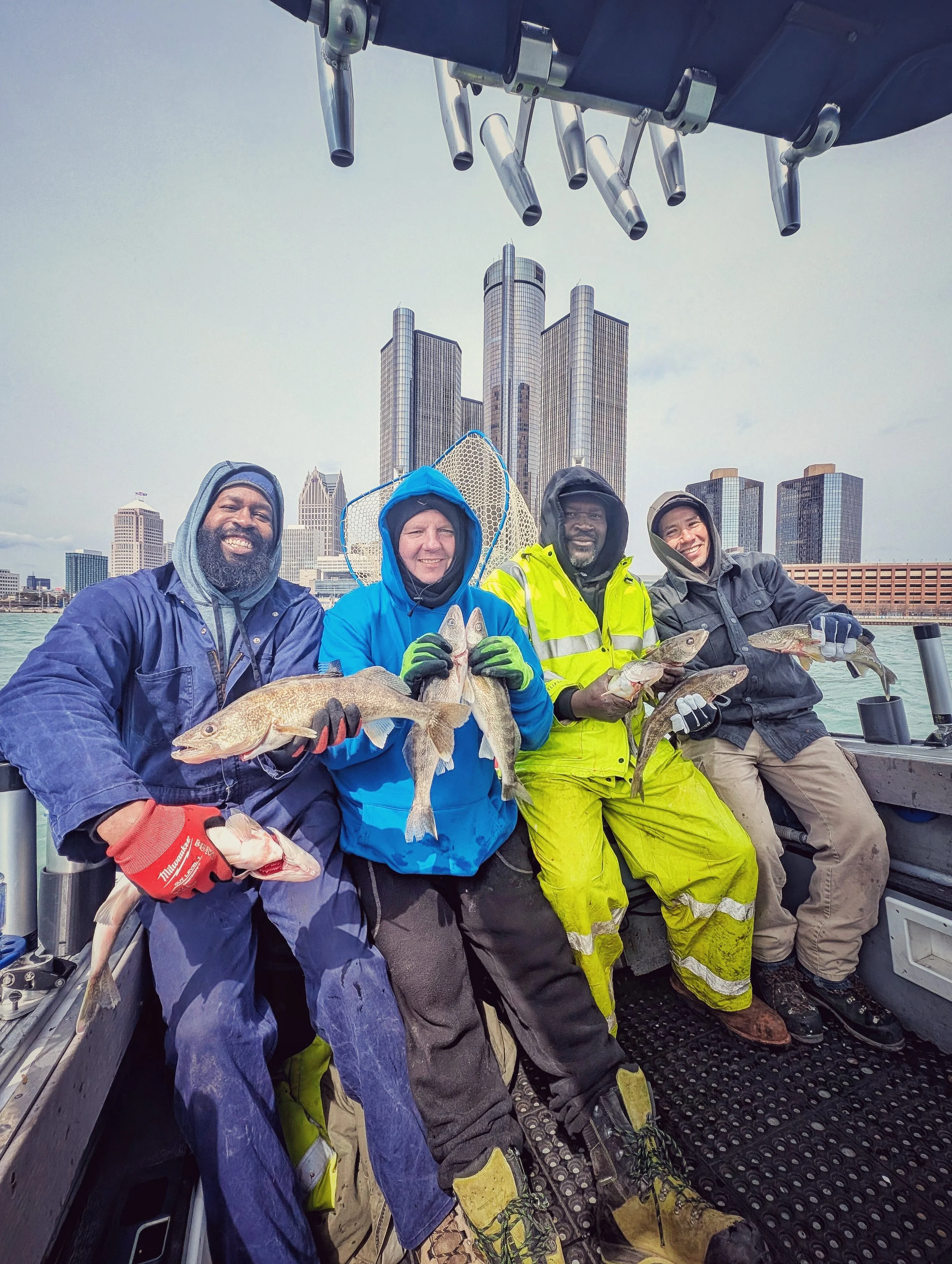  A diverse group of four anglers in rain gear holding up walleye on a Reel Fish'n charter boat with the Detroit Renaissance Center skyline in the background. 
