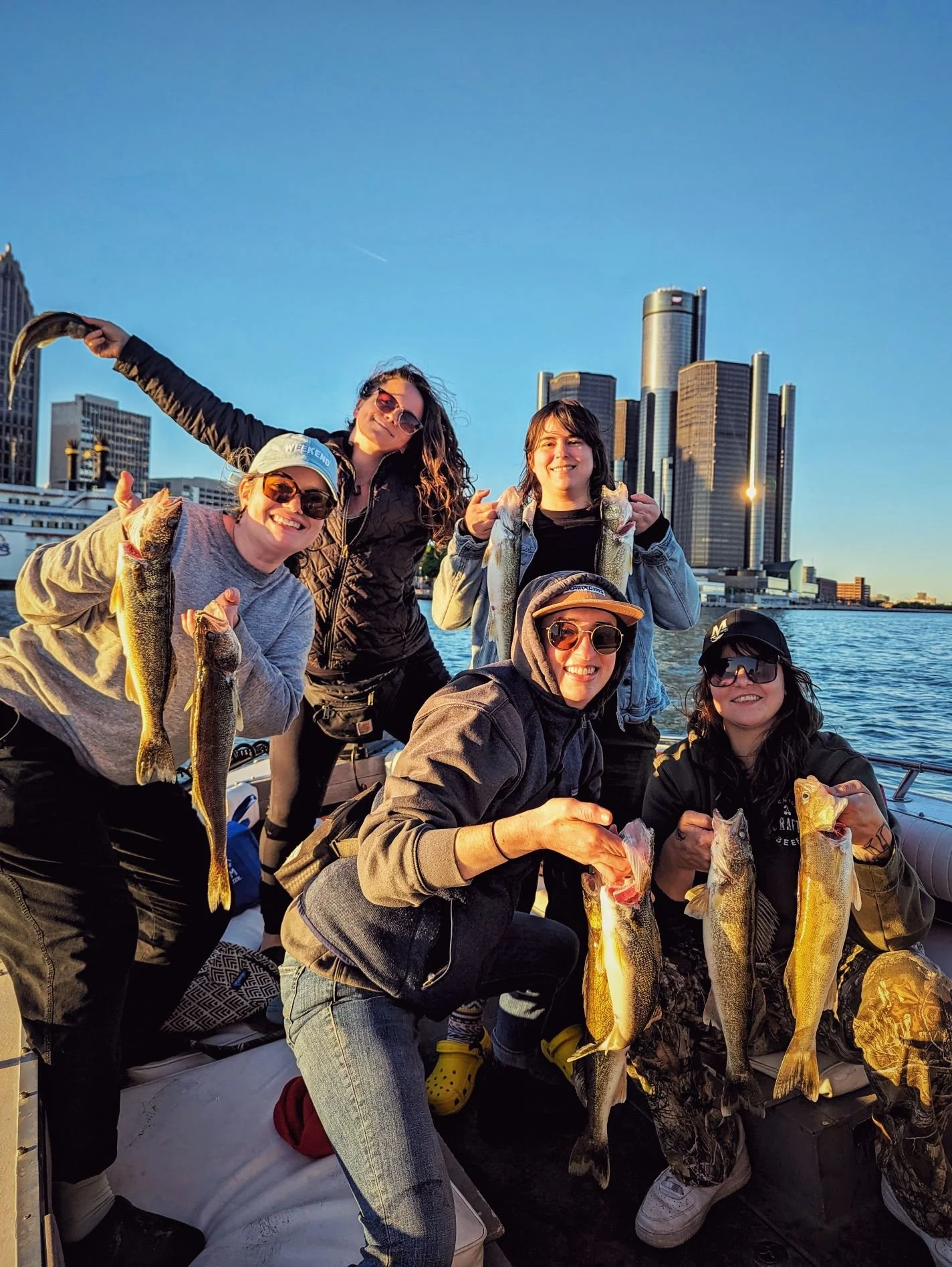  A group of women holding up their walleye catch with the Detroit Renaissance Center and city skyline in the background during a sunset charter.   