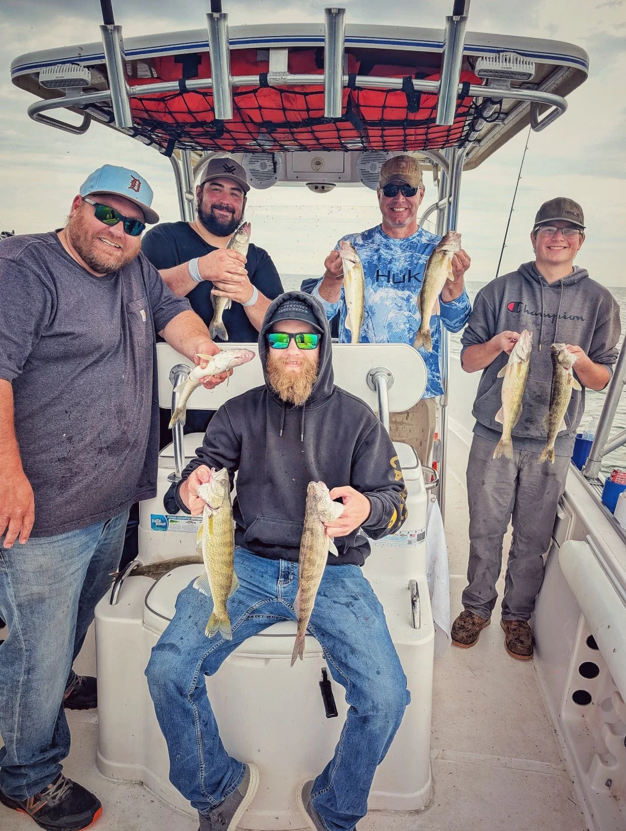 Group of five men on a boat holding caught fish, smiling. Four men hold fish in their hands, and one man holds two fish, sitting on a cooler, in a cloudy sky background.