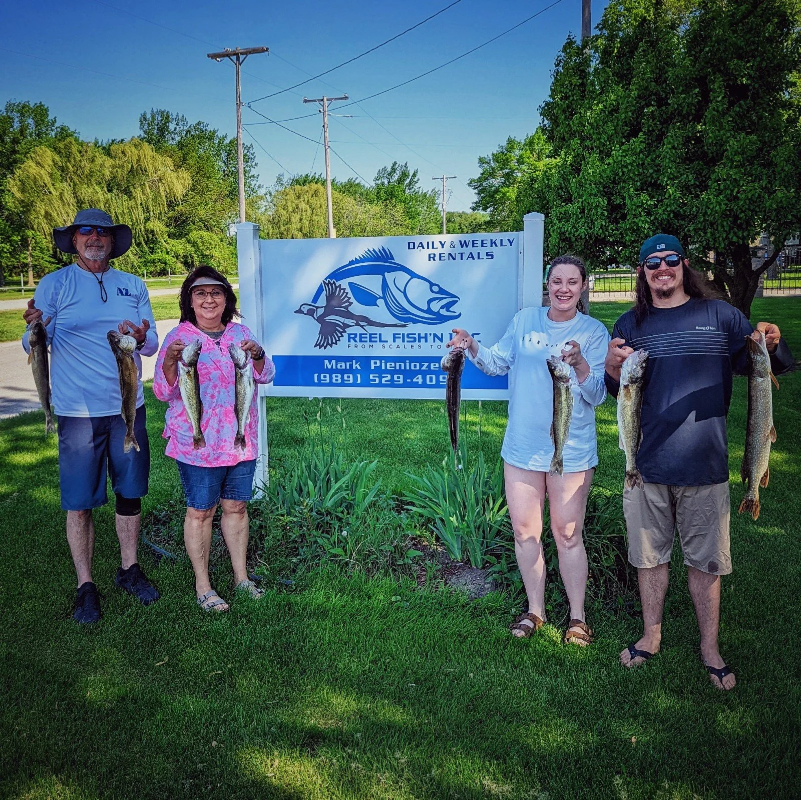 A group of five people standing outdoors holding fish, posing in front of a sign that reads 'Reel Fish'N From Scales To Catch' and 'Daily & Weekly Rentals', during a sunny day with green trees and a blue sky in the background.