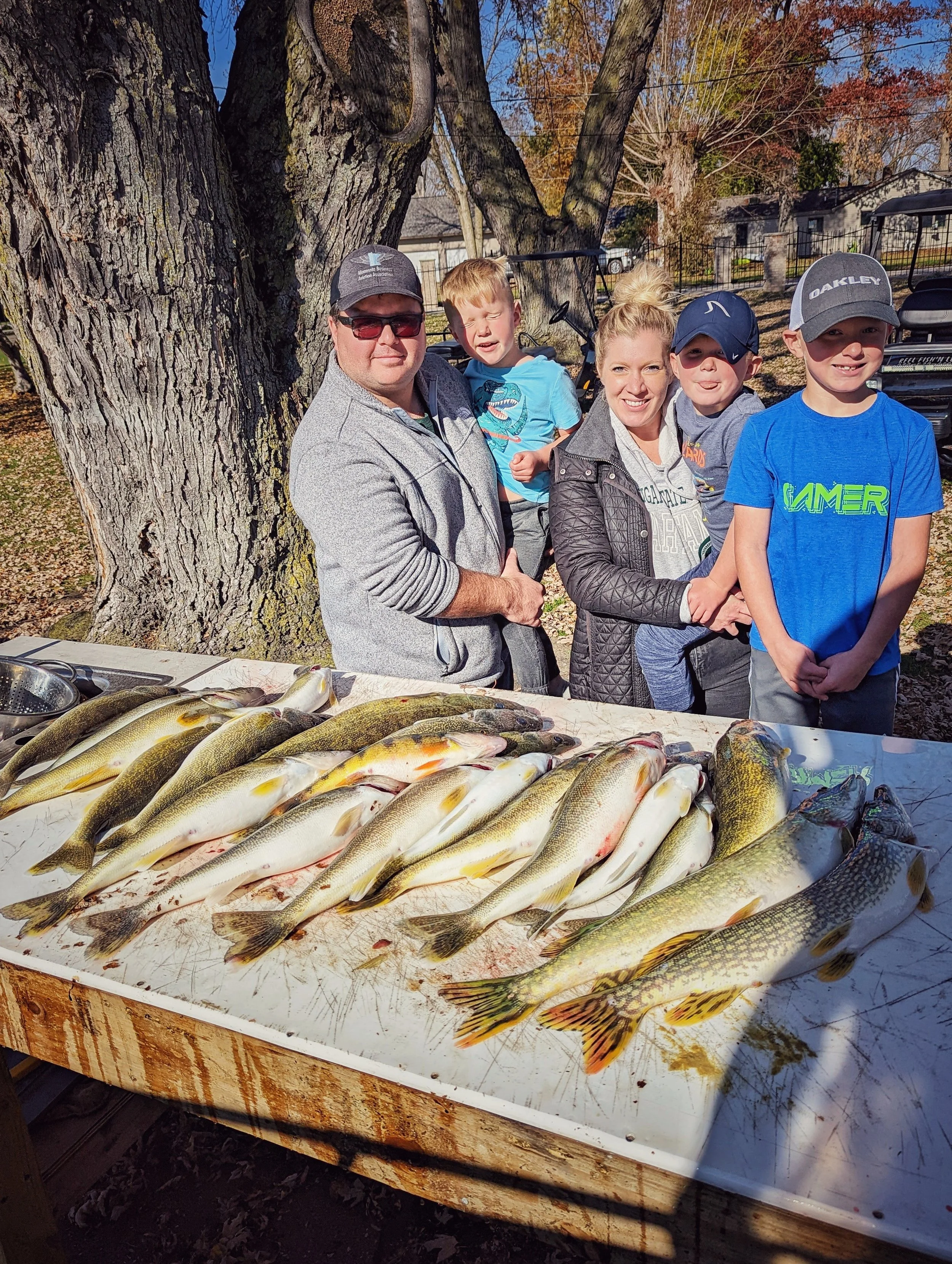 A group of five people, a man, a woman, and three boys, standing behind a table with caught fish, outdoors near large trees with autumn foliage.