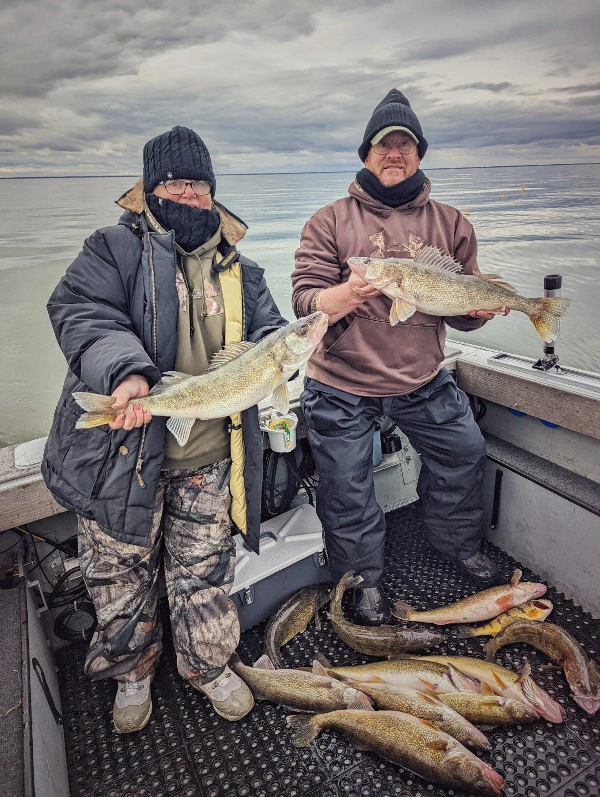 Two men on a boat holding large fish they caught, with several fish lying on the boat's floor, against a body of water and cloudy sky.