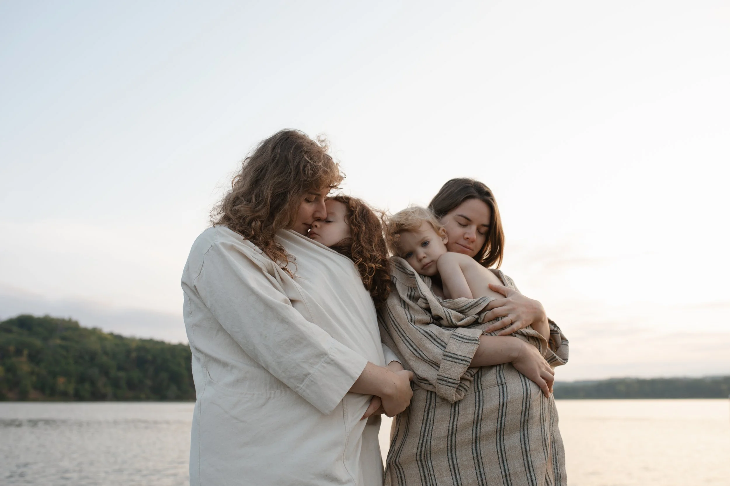 Three women holding children during sunset near a river, with a green hillside in the background.