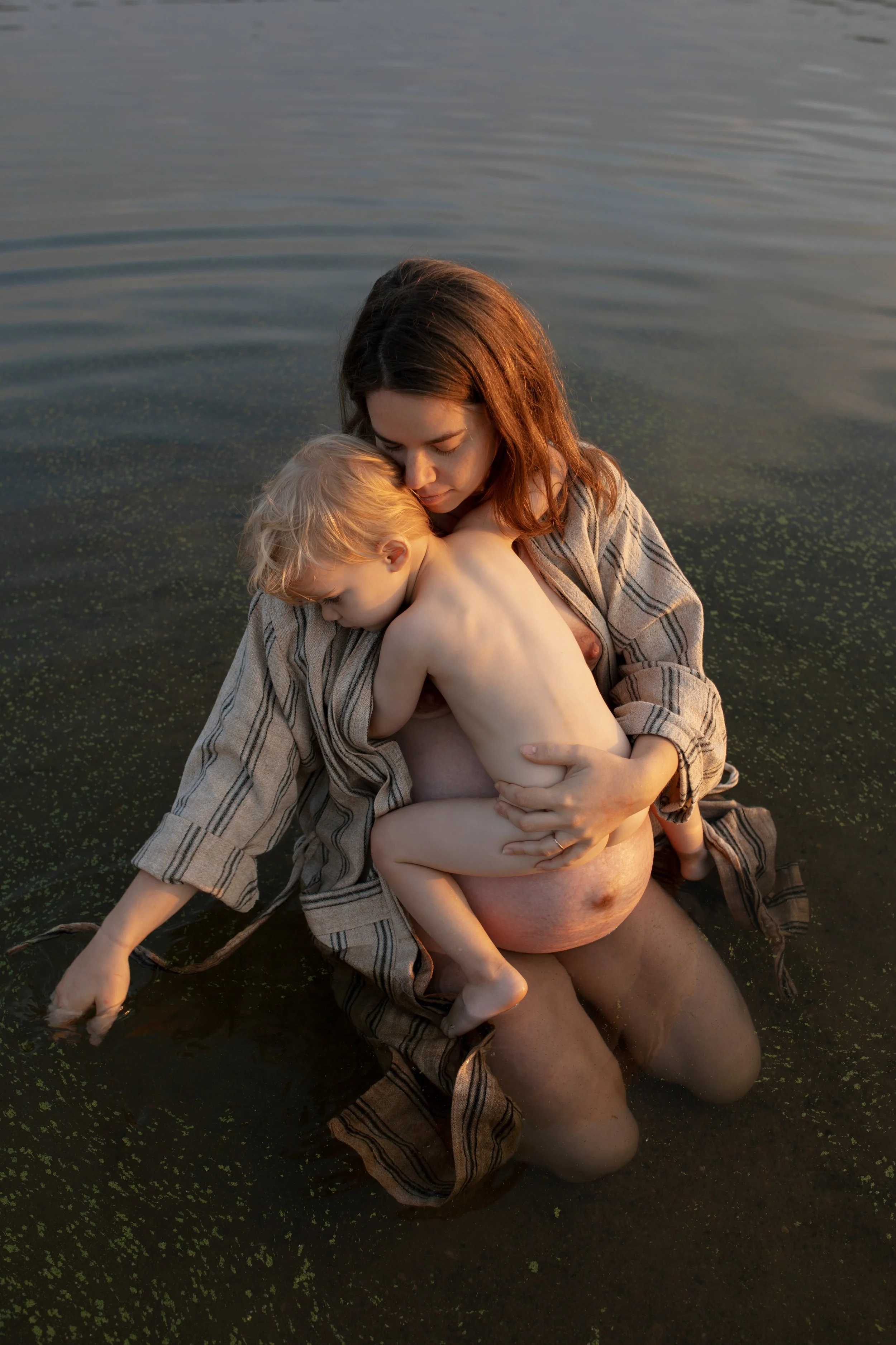 A woman and a young boy in a lake during sunset, with the woman holding the boy close as they sit in the water.