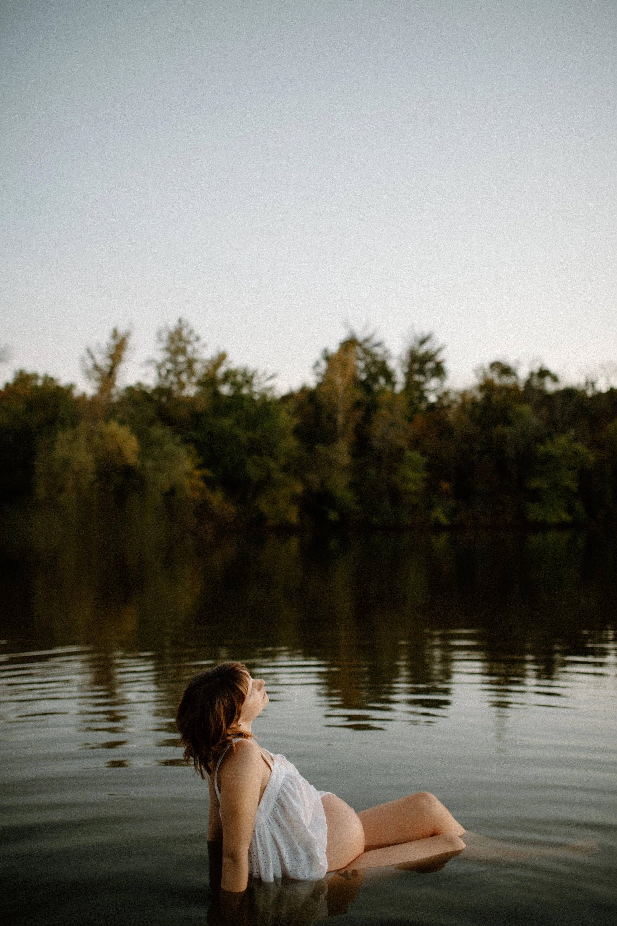 A pregnant woman with brown hair wearing a white dress reclining in a calm lake surrounded by trees, during sunset.