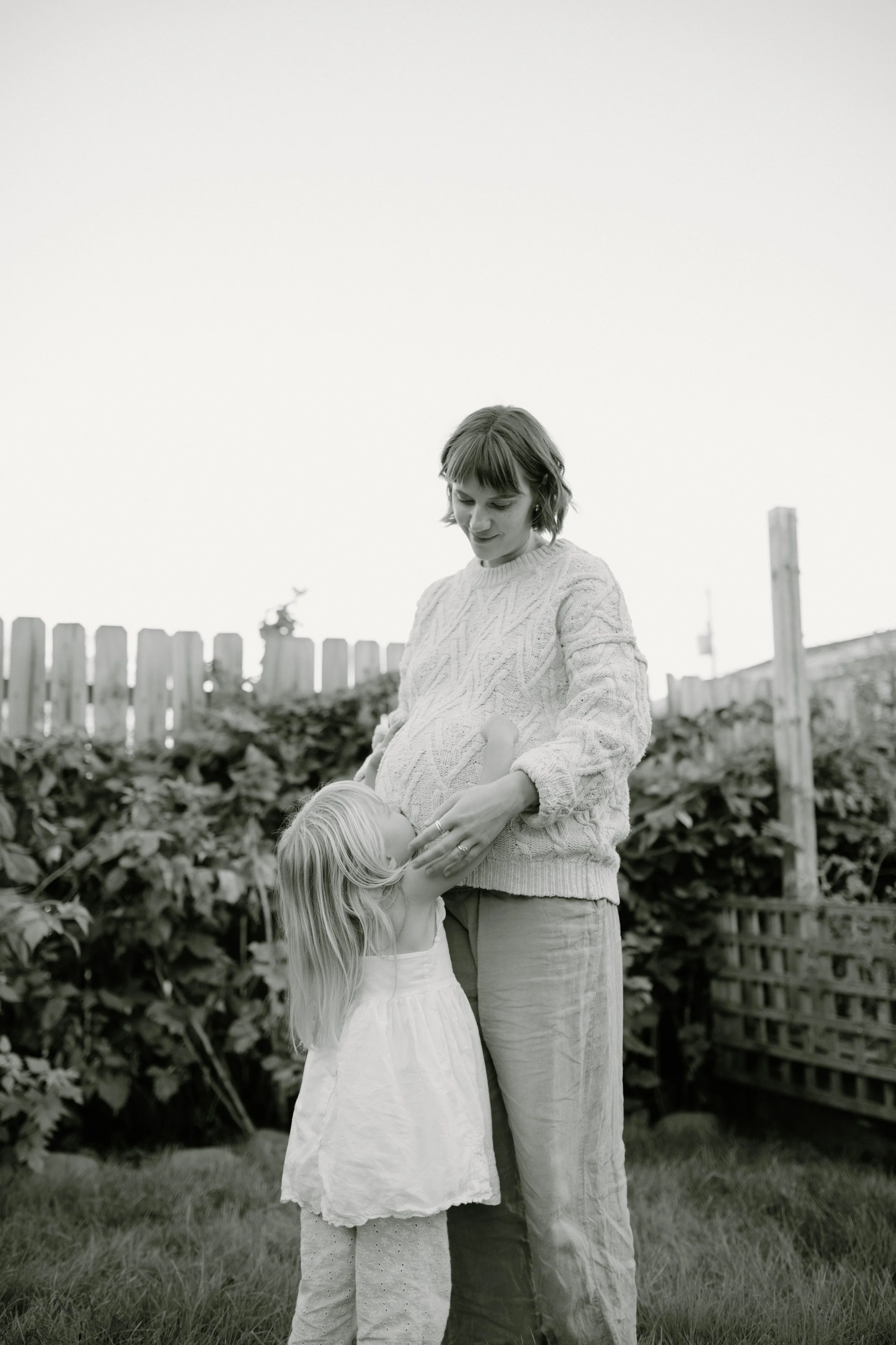 A woman with short hair and a girl with long hair standing outdoors in a garden or yard, with a wooden fence and plants in the background. The woman appears to be helping or caring for the girl.