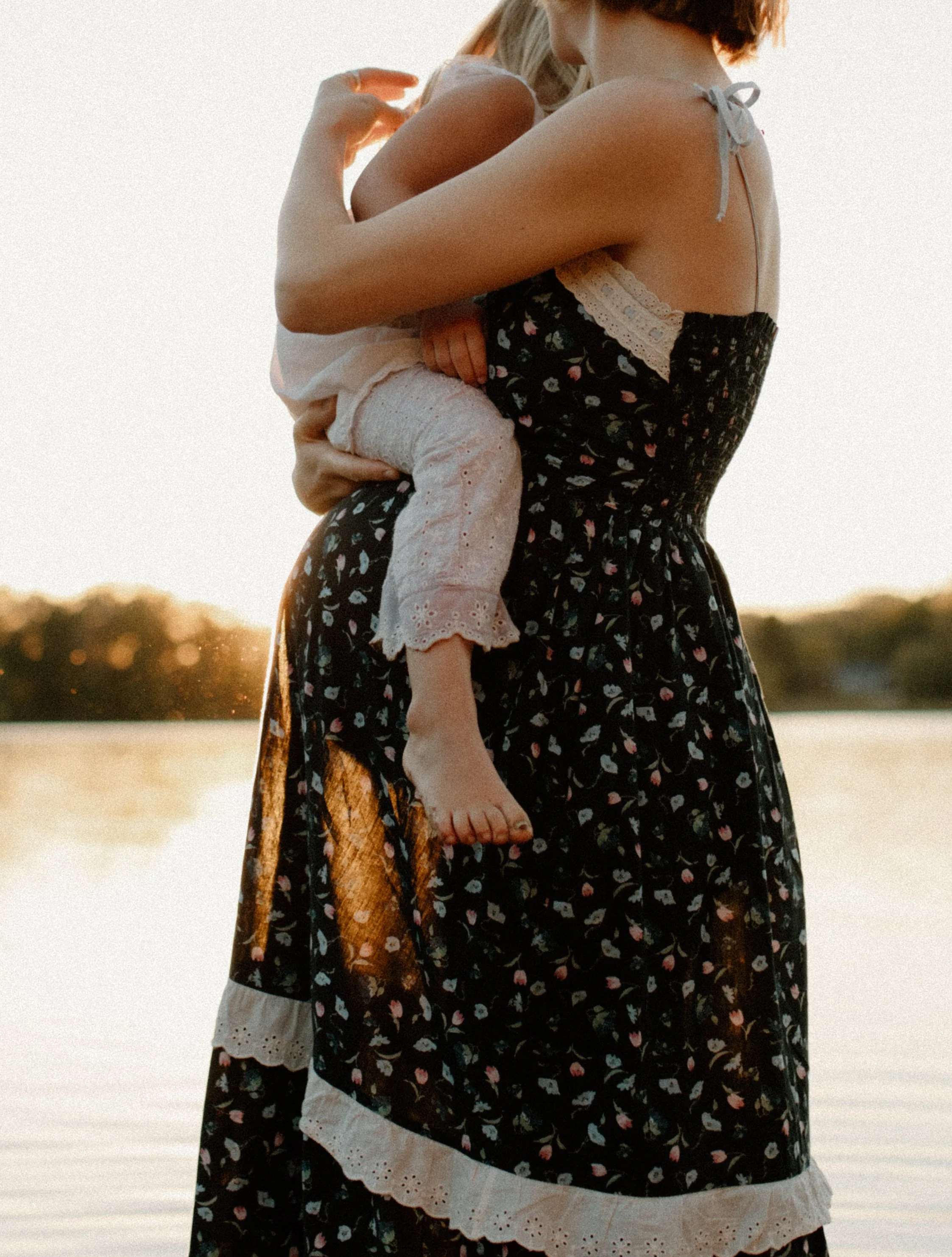 A pregnant woman holding a small child near a body of water at sunset.