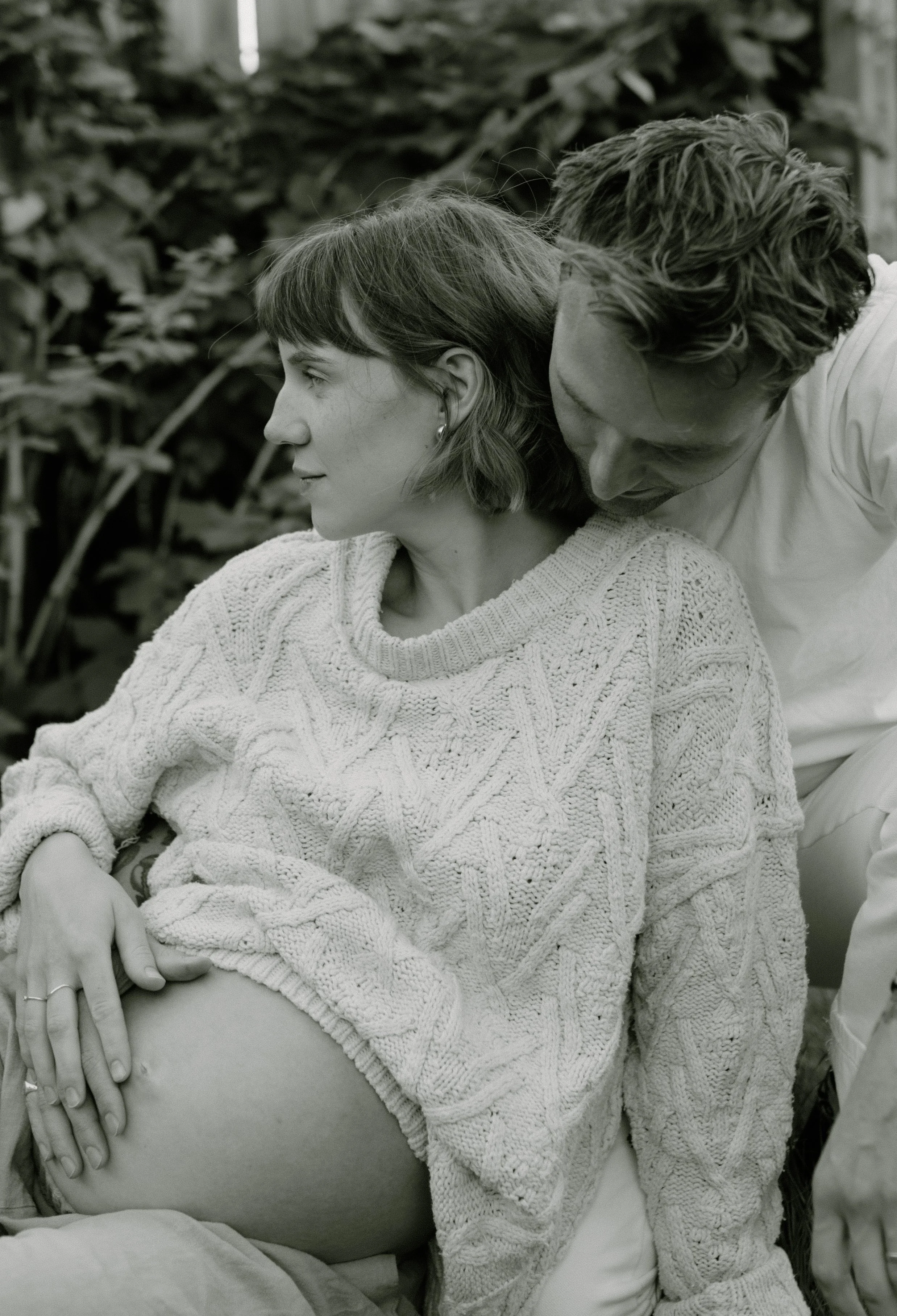 A woman with short hair and earrings, wearing a knitted sweater, sitting with a man leaning over her, with his face close to her neck, outside in front of a backdrop of leafy plants.