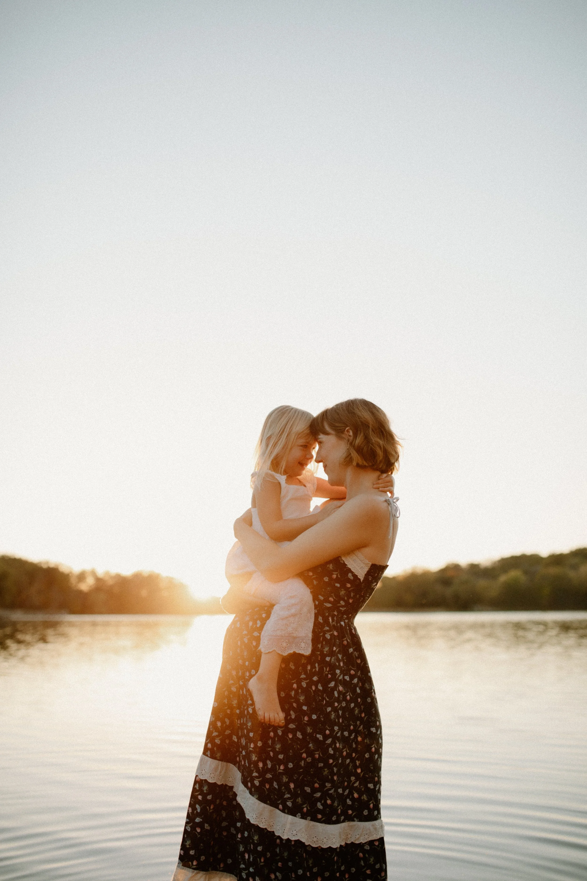 A woman holding a young girl by a lake during sunset. The woman is wearing a black dress with a white scalloped hem, and the girl is in a white dress. They are touching foreheads and smiling with their eyes closed.