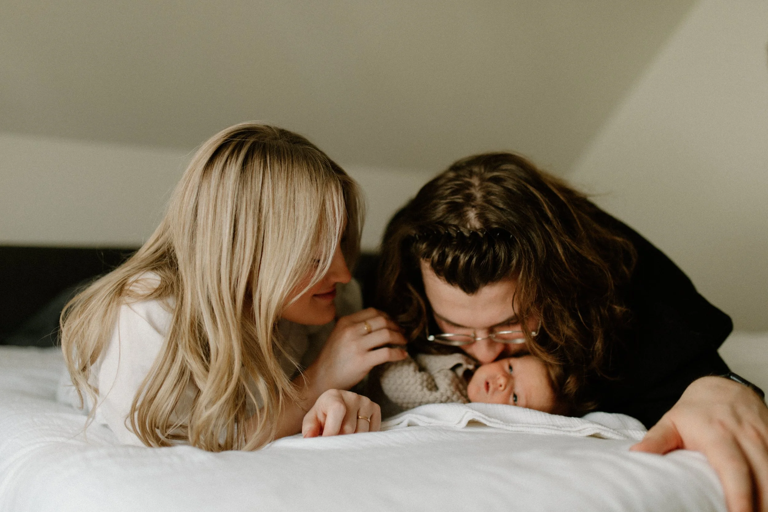 A young couple leaning over a bed, smiling and holding a newborn baby.