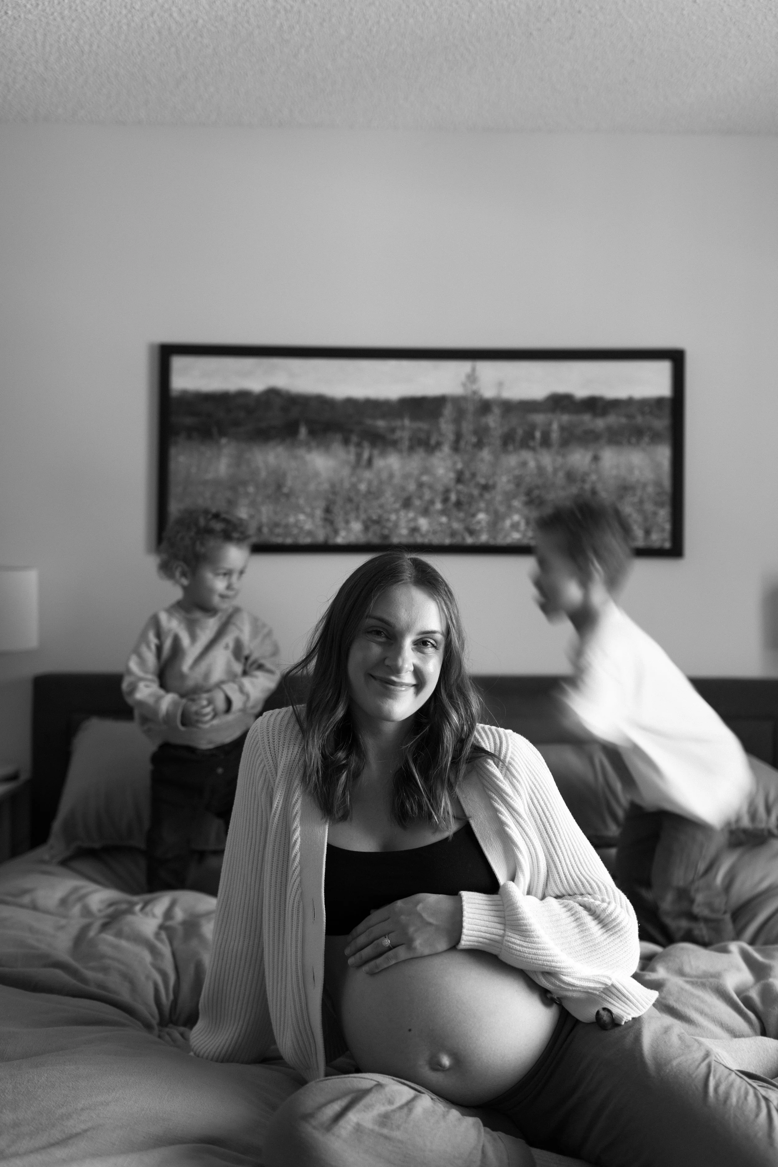 A woman with long hair sitting on a bed with a pregnant belly, smiling at the camera, with two young boys in the background in a bedroom with a landscape painting on the wall.