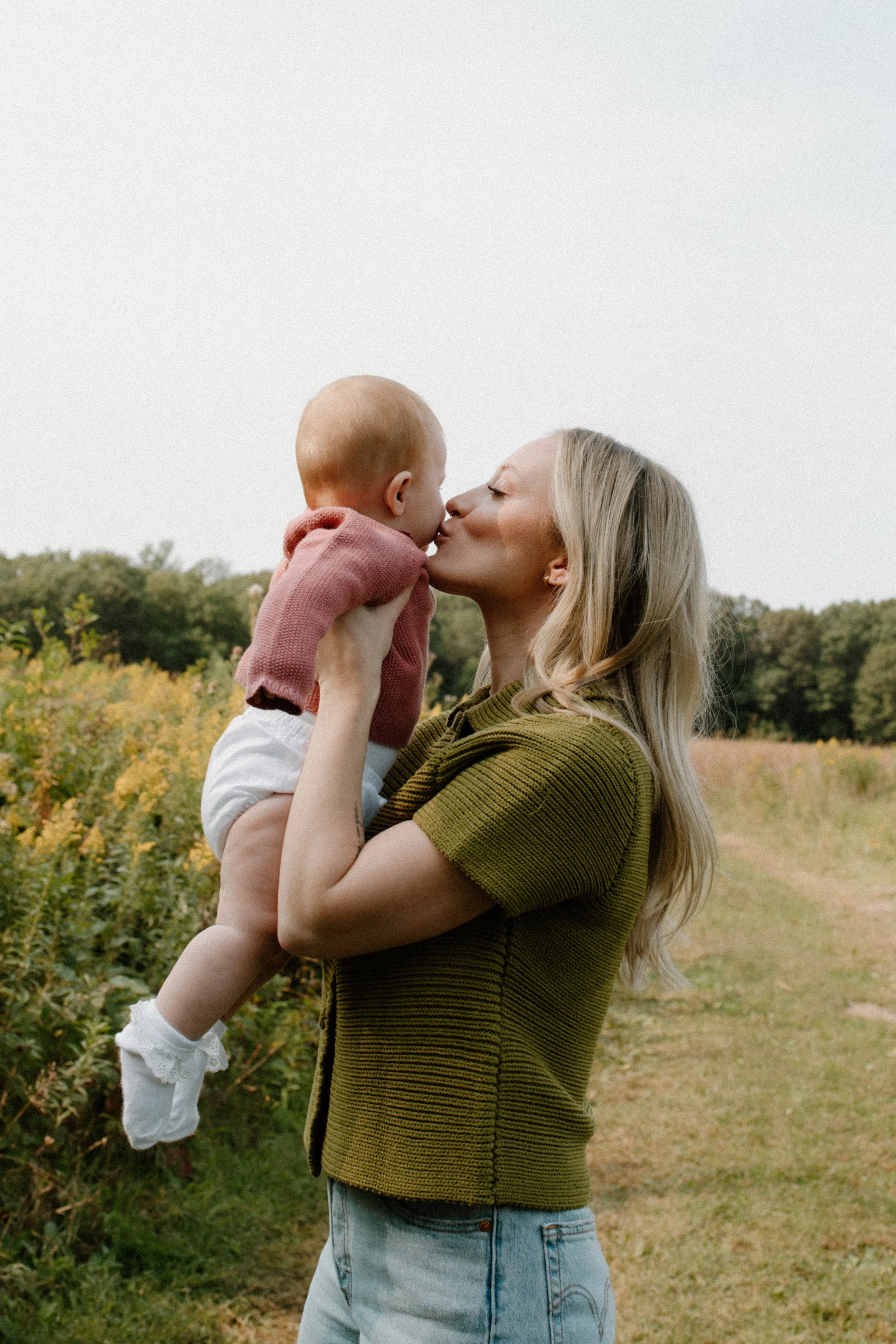A woman holding a young child, they are kissing outdoors in a field with greenery and fall foliage in the background.