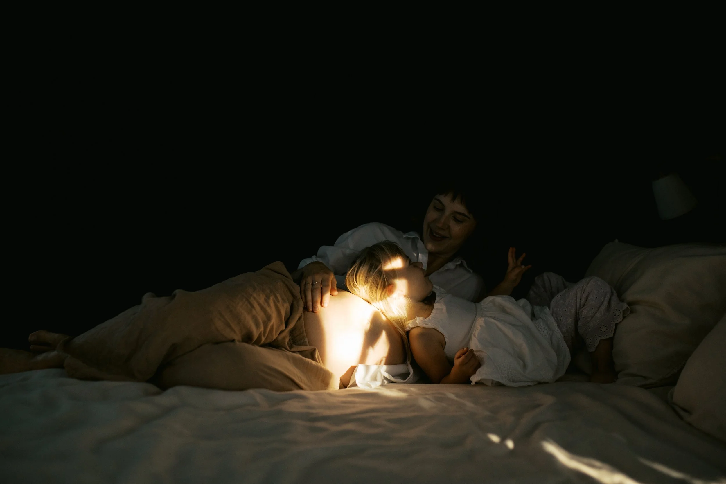 A woman and two children lying in bed at night, with one child holding a flashlight and the woman smiling.