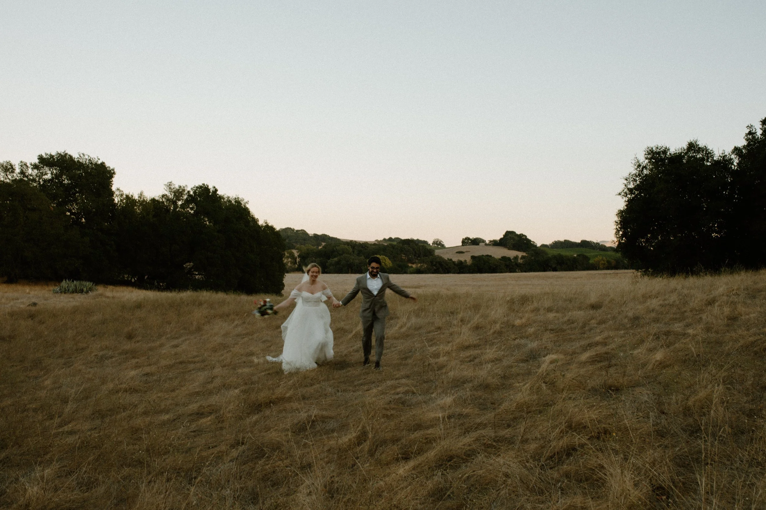 A bride and groom walking hand in hand across a field at sunset, with trees and rolling hills in the background.