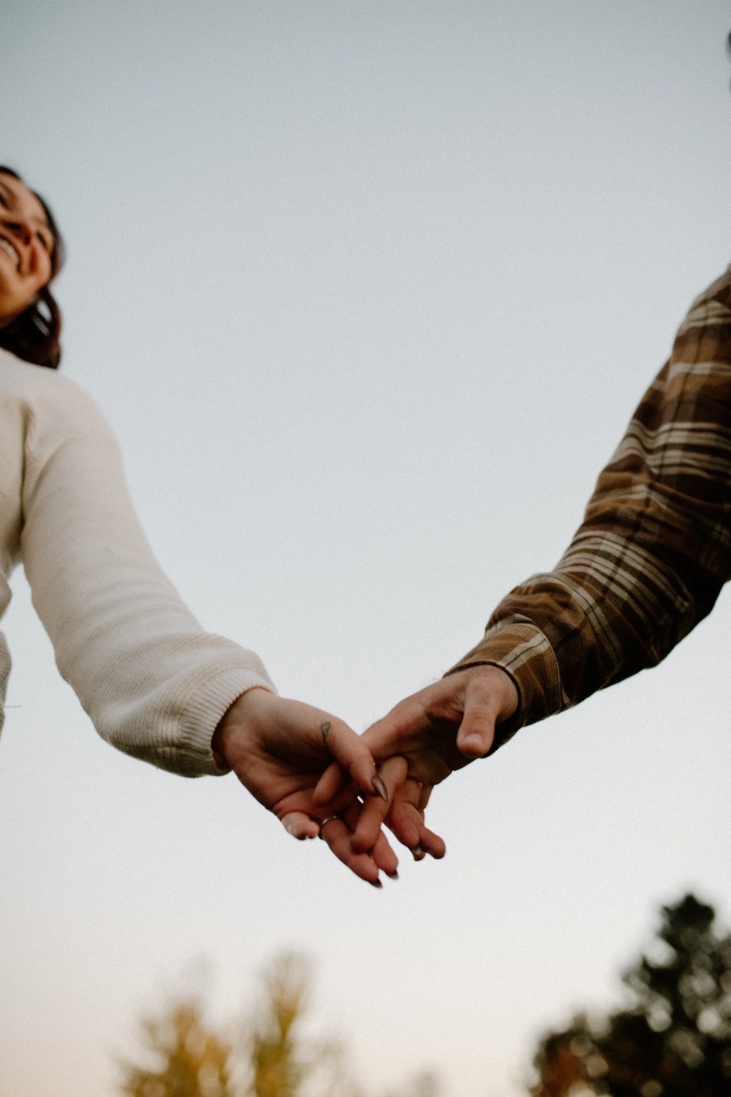 Two people holding hands with fingers interlocked, visible from their arms and hands, against an outdoor background with trees and sky.