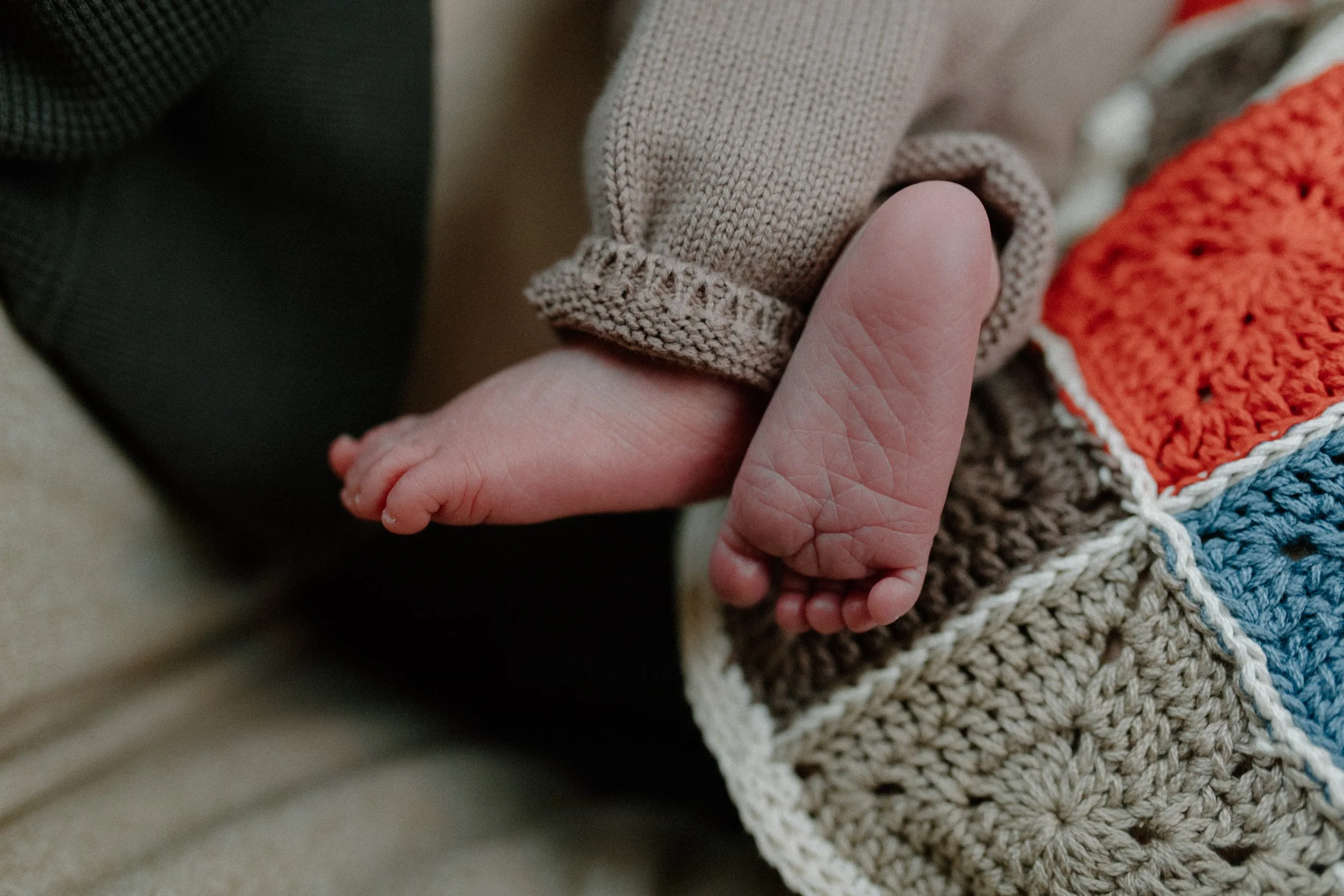 Close-up of an adult hand gently holding a small baby's hand, with the baby wearing a beige knitted sleeve, and part of a colorful knitted blanket visible.