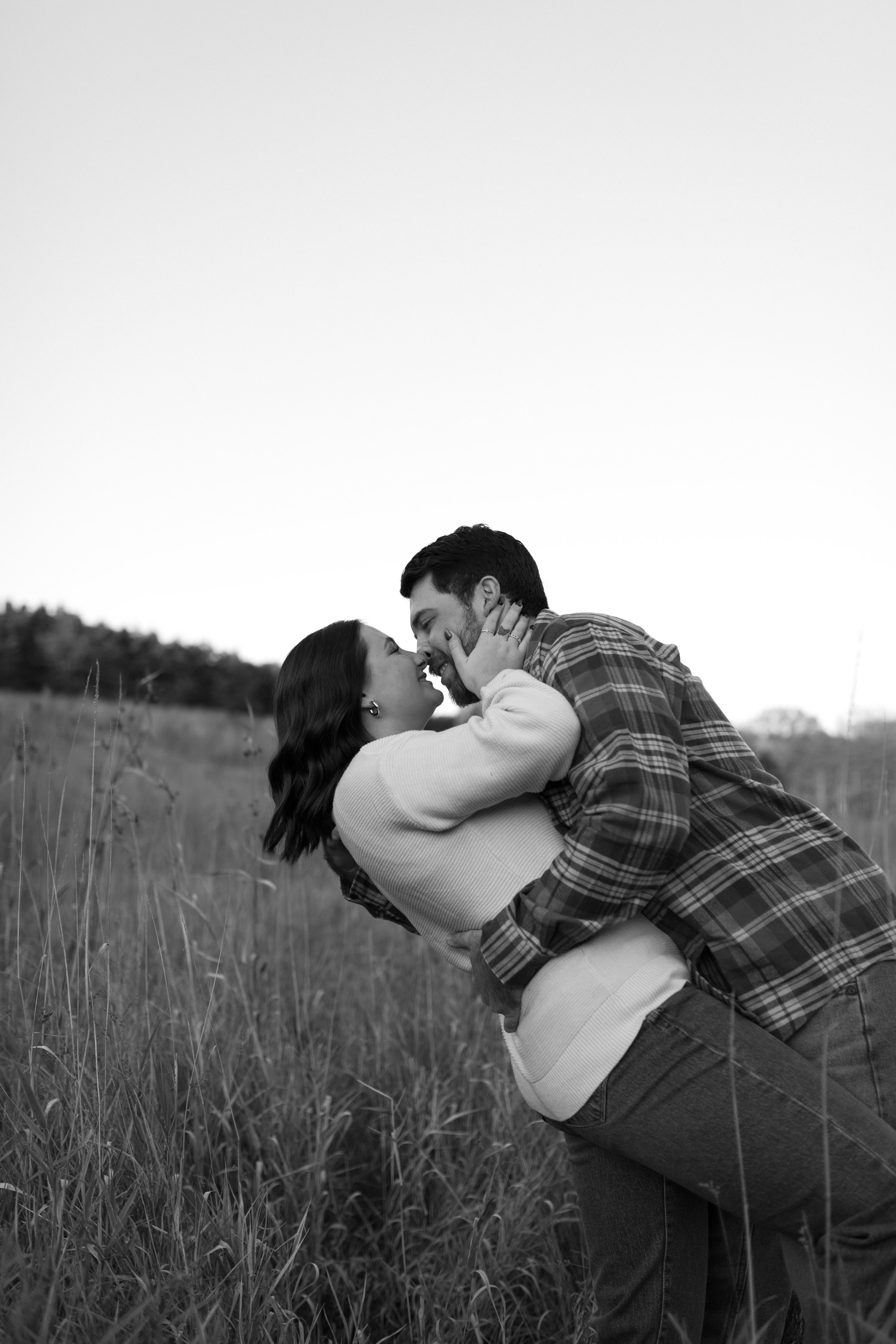 A couple embraces and shares a kiss in a grassy outdoor field with a slightly blurred treeline in the background.