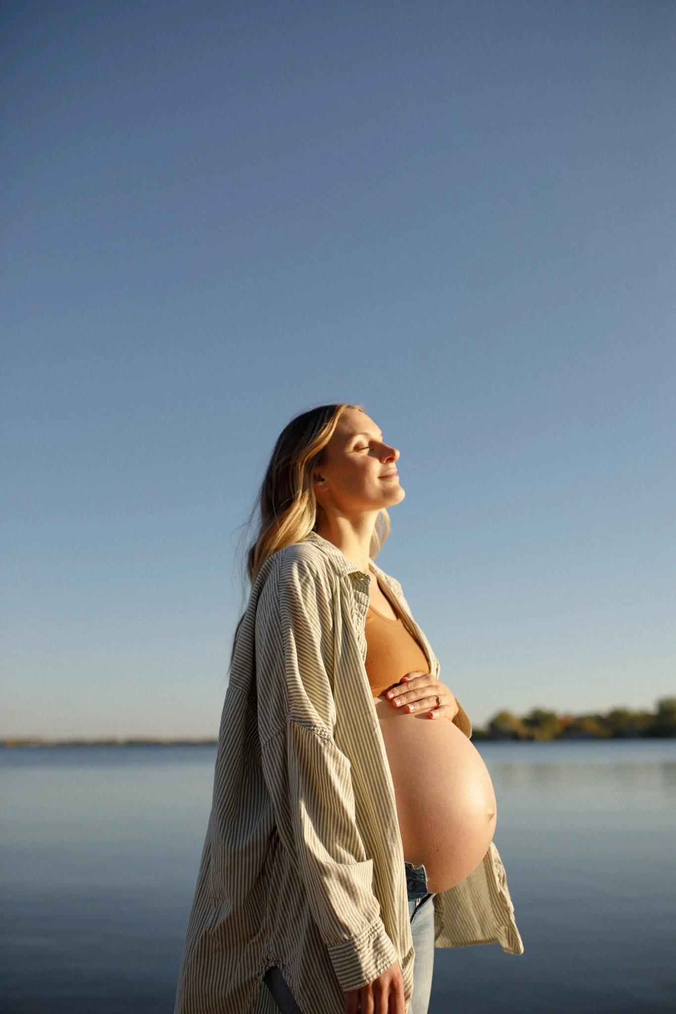 A pregnant woman with blonde hair standing outdoors by a body of water, with her eyes closed and a peaceful expression on her face, wearing a beige open shirt over her bare belly, during what appears to be sunset or sunrise.