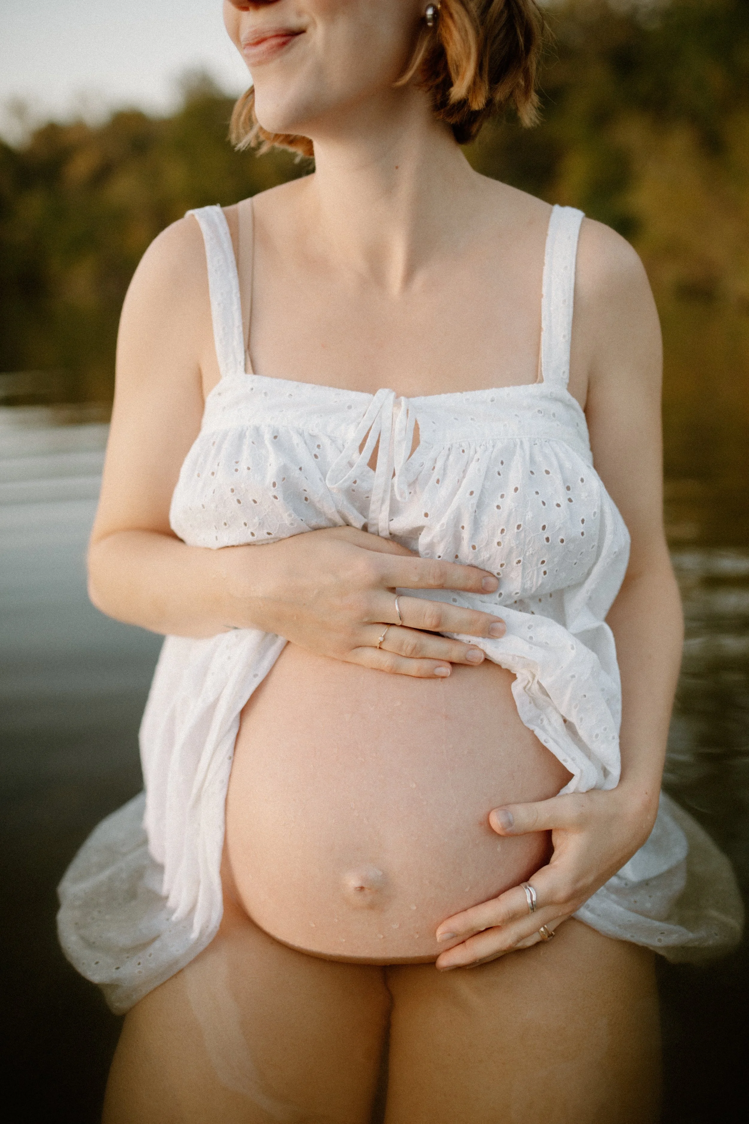 A pregnant woman is standing outdoors near a body of water, wearing a white eyelet dress with thin straps, with her hand on her abdomen and a slight smile on her face.