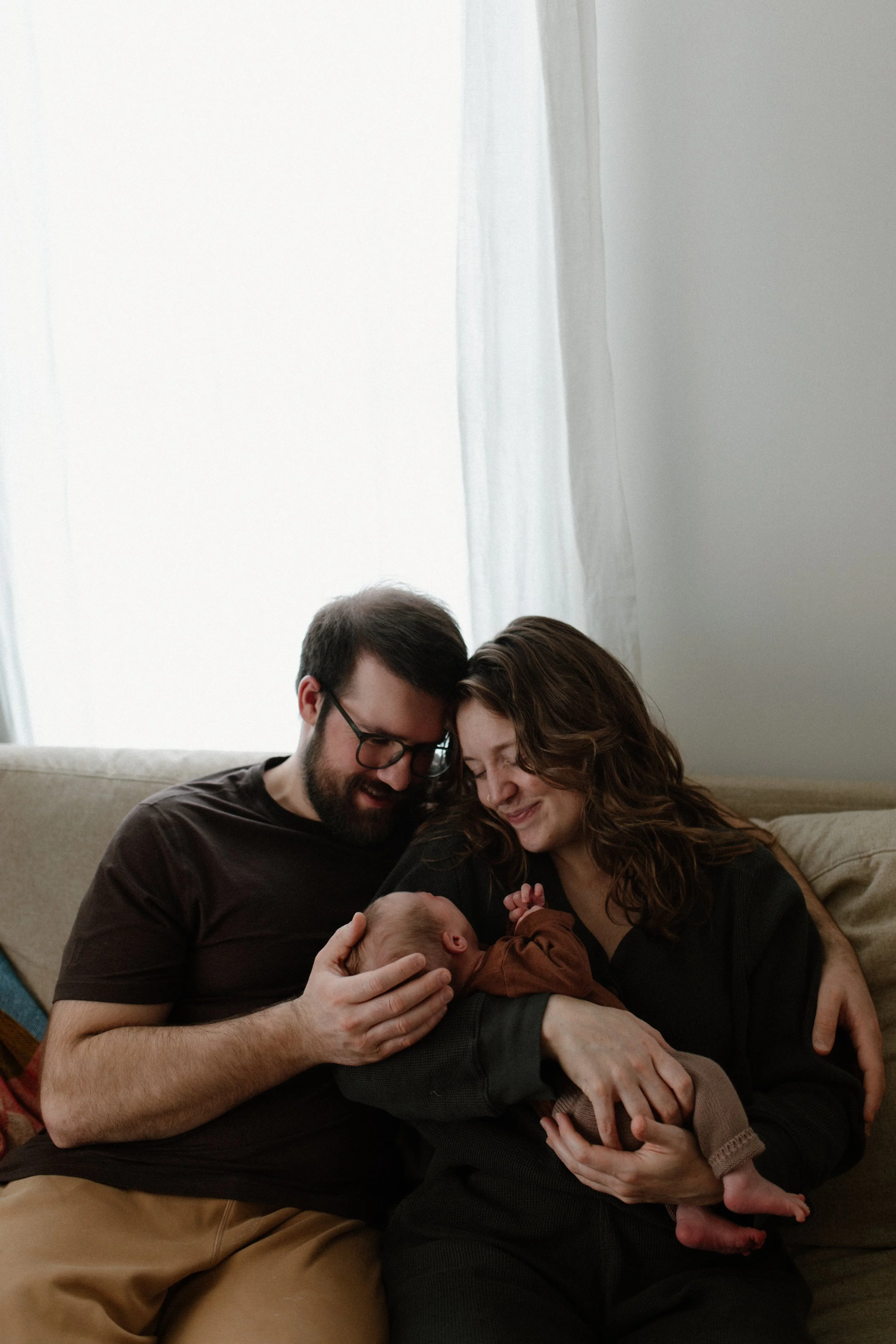 A family of three, including two parents and a newborn baby, sitting on a couch indoors, smiling and bonding with the baby.