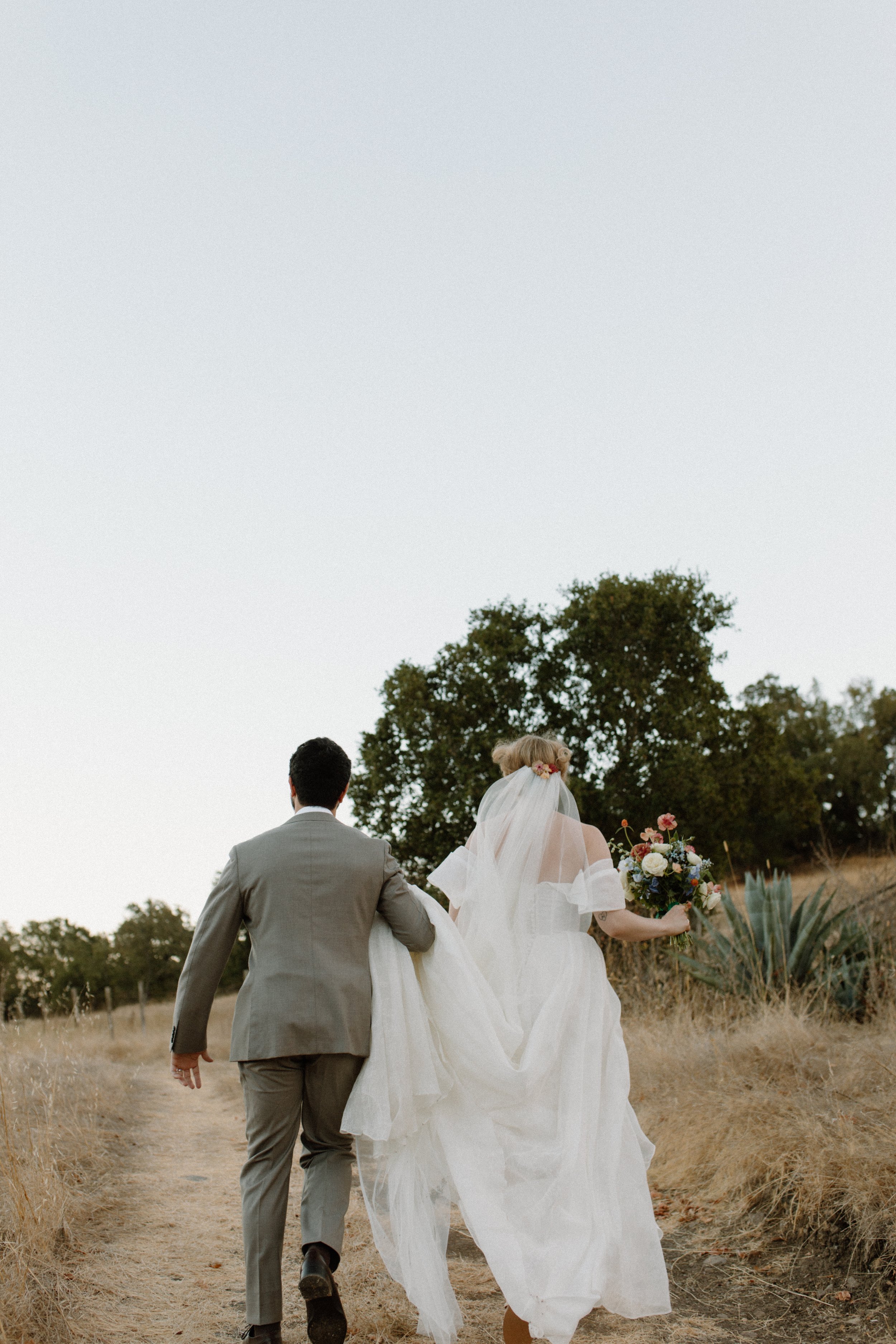 A bride and groom walking away together outdoors on a dirt trail, with the bride holding a bouquet, during daytime.