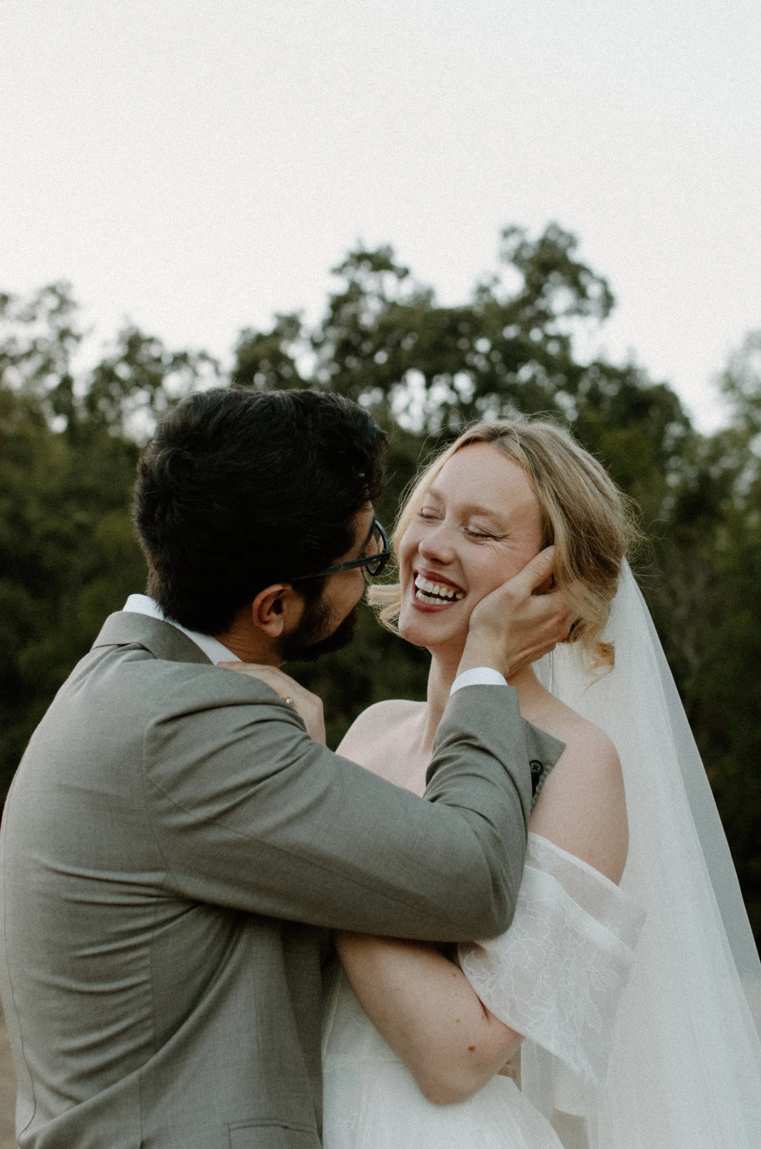 A couple, with the man wearing a gray suit and glasses, and the woman in a white wedding dress, sharing an intimate moment outdoors during their wedding, with trees and an overcast sky in the background.
