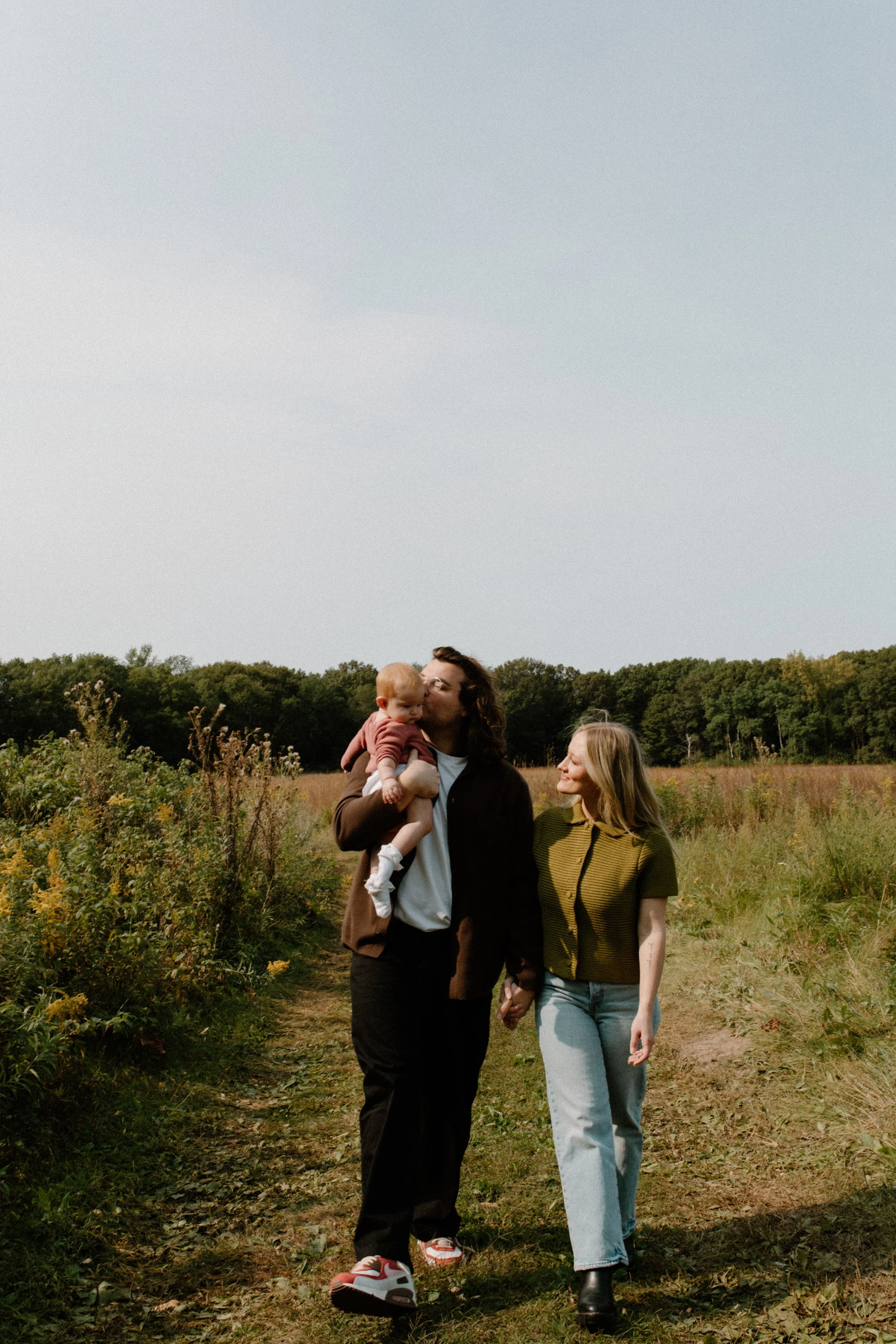A family of four holding hands walks through a grassy field with trees in the background during daytime. The father, holding a baby, kisses a woman, while a girl walks beside them.