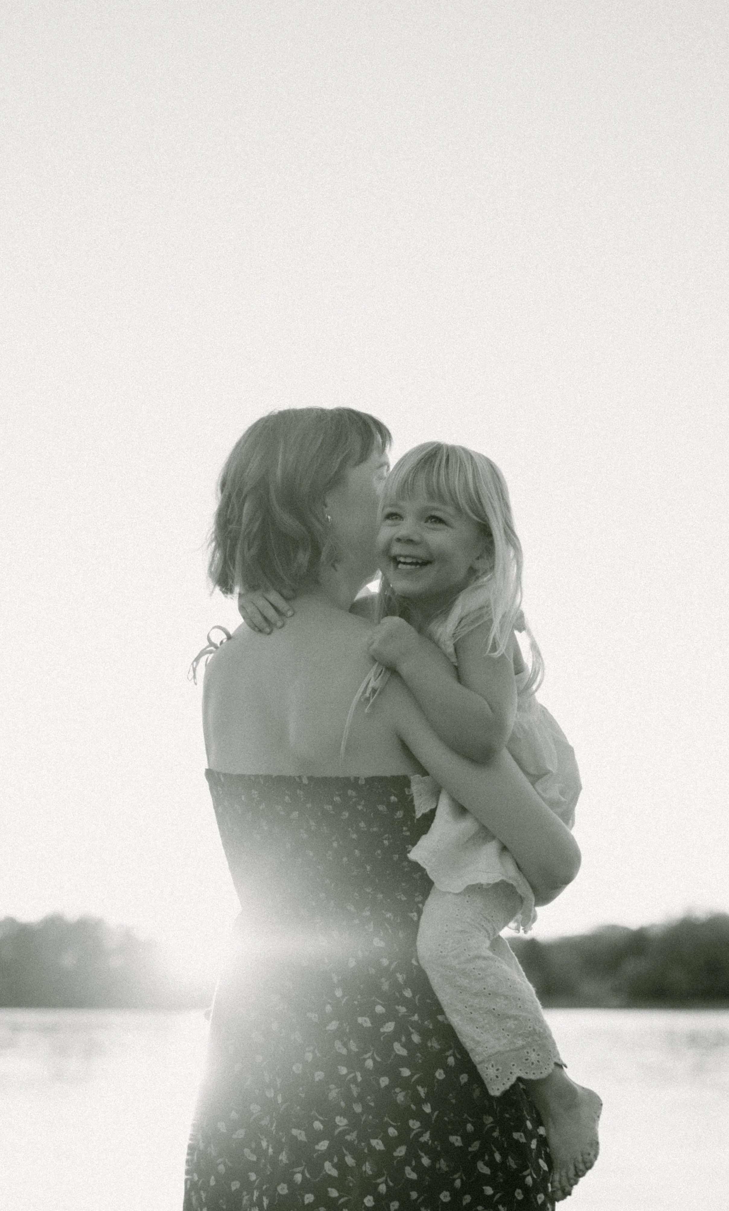 A woman holding a smiling young girl in her arms outdoors near a body of water, with trees in the distance, in black and white.