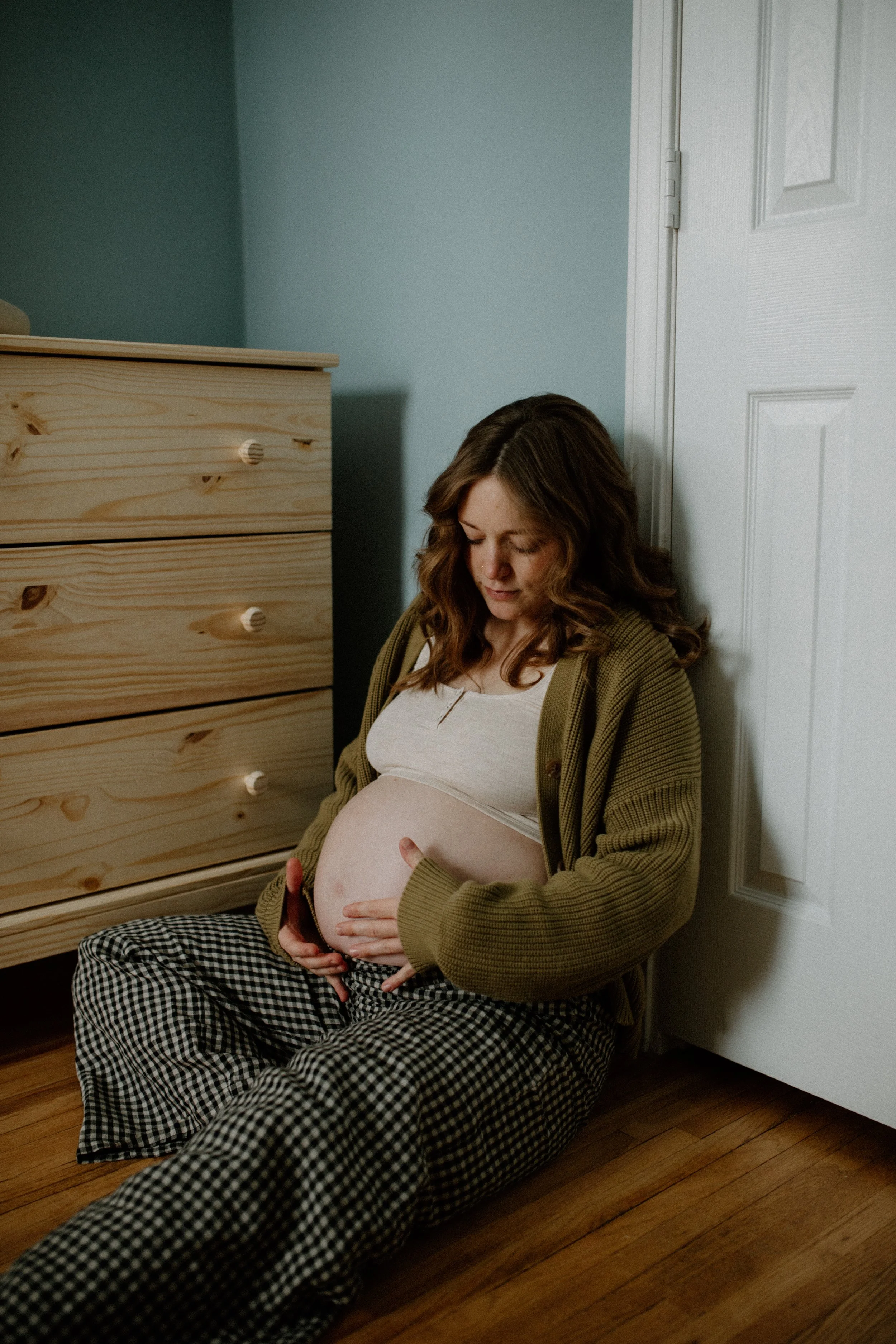 Pregnant woman sitting on the floor, leaning against a wall, with one hand on her belly and the other hand holding her phone, near a wooden dresser in a room with hardwood flooring.