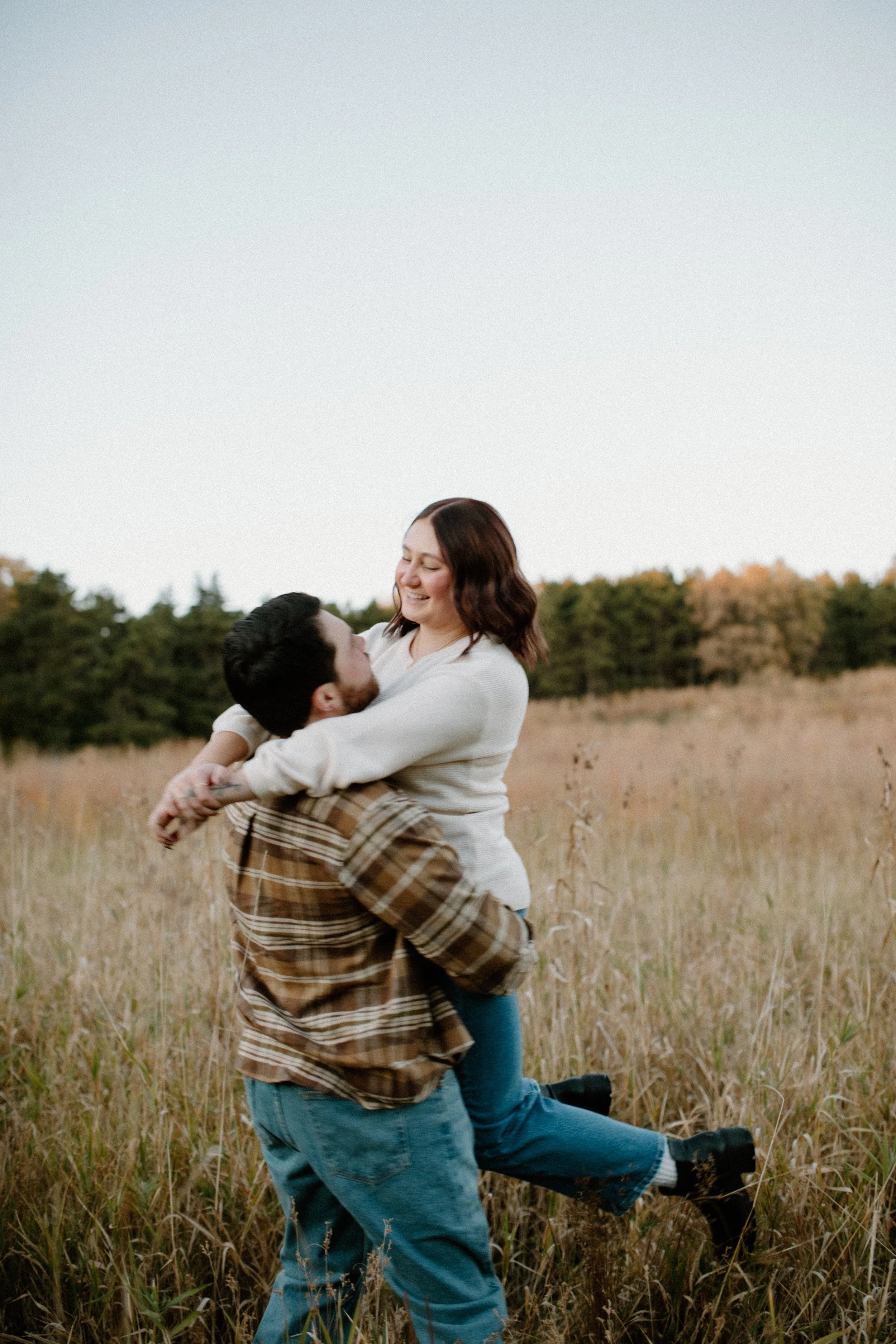 A woman holding a man in a field of tall grass, smiling, with a backdrop of trees and a clear sky.