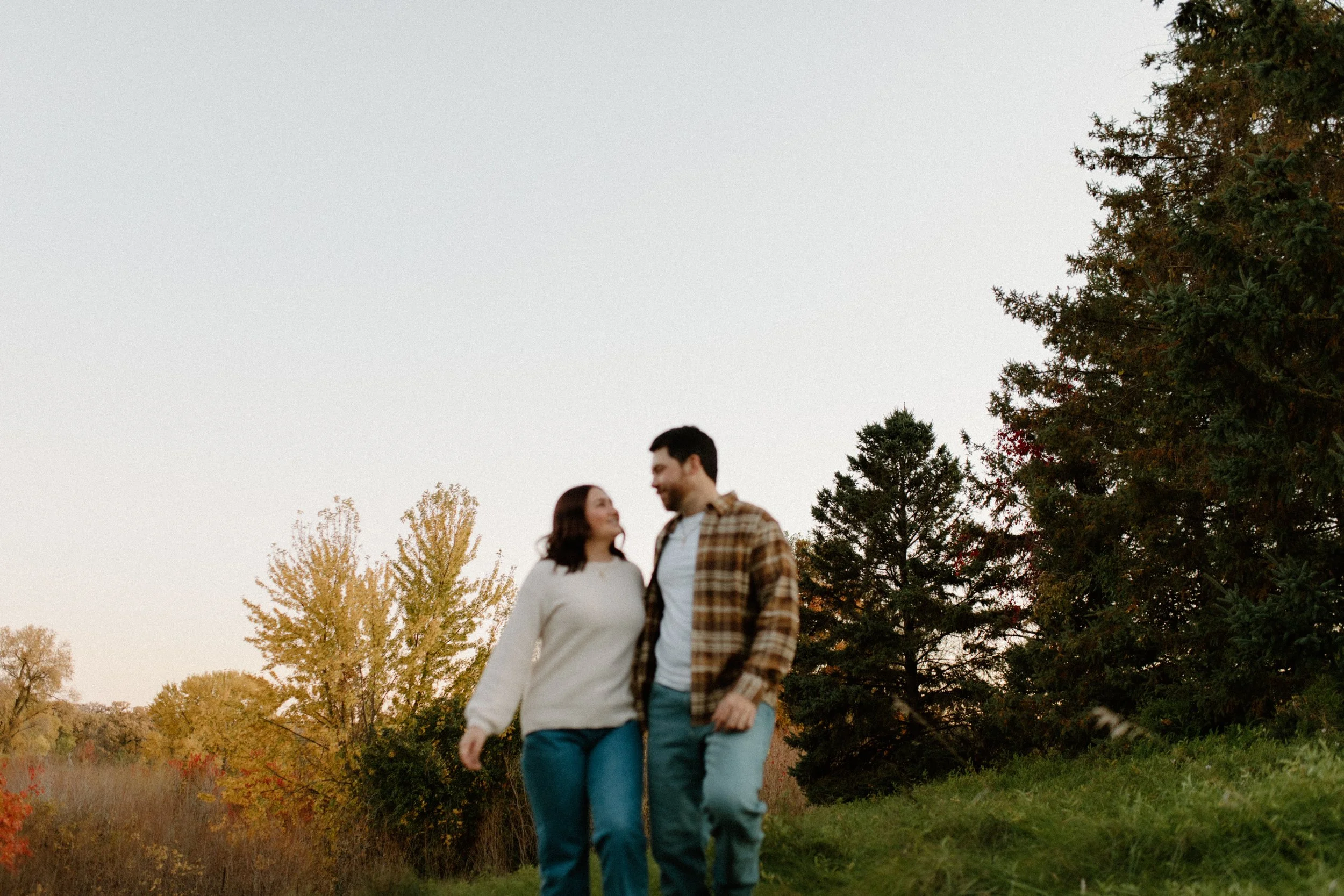 A couple walking outdoors in a park during autumn, with trees displaying fall foliage around them.