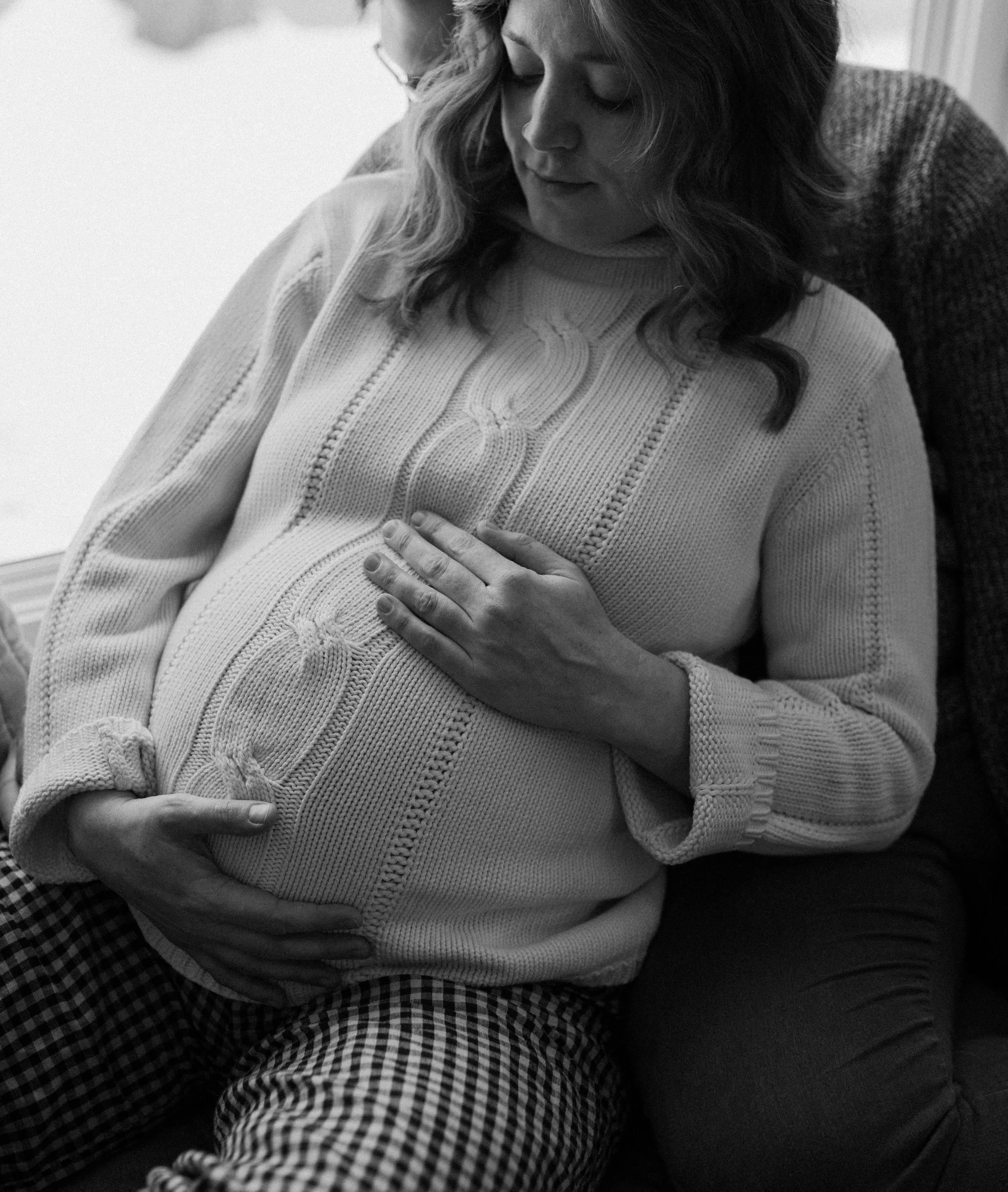 A woman with long wavy hair is seated, gently holding her stomach with her eyes closed, indicating she might be feeling unwell or experiencing discomfort.