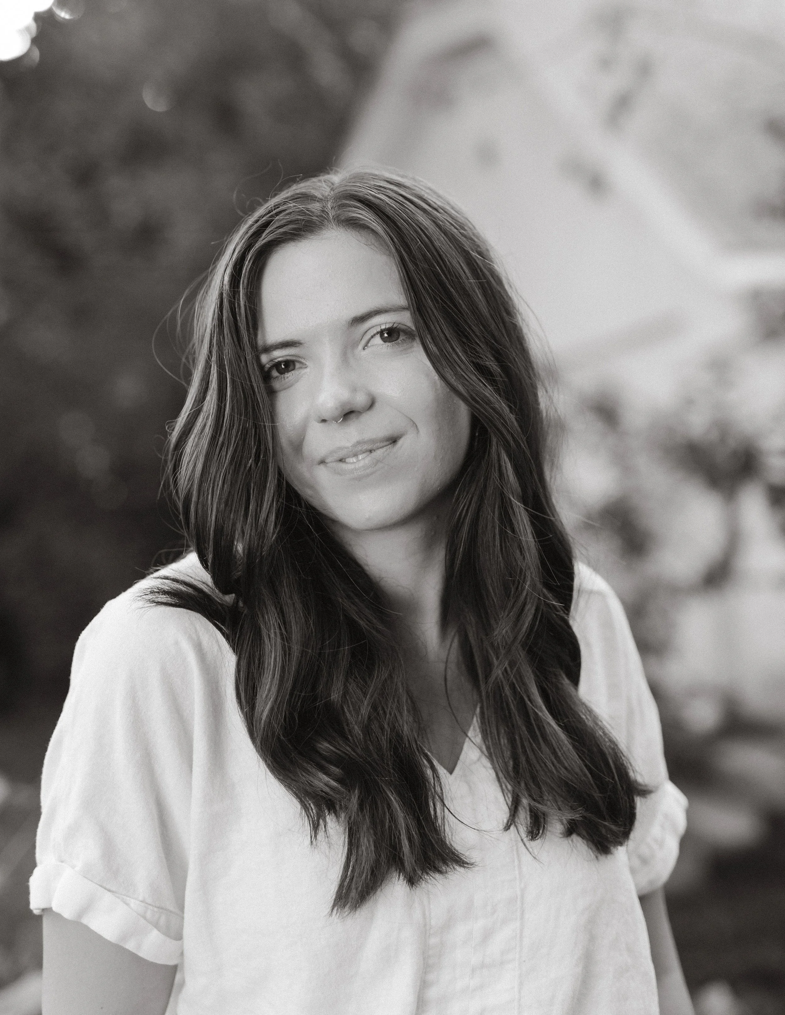 Black and white photo of a woman with long, wavy hair and a septum piercing, smiling softly, wearing a light-colored top, outdoors with blurred background