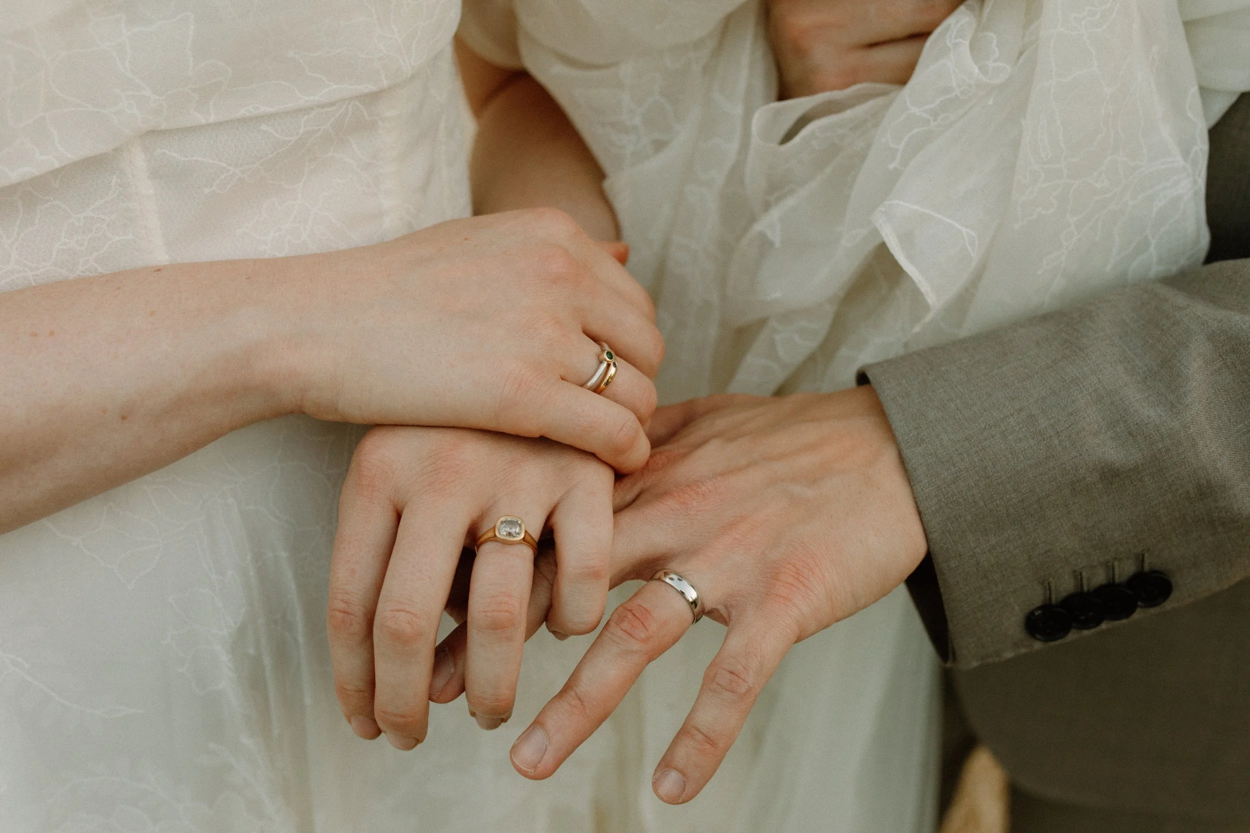 Close-up of a couple's hands as they hold each other, showing wedding rings on both their fingers. The bride's hand has a gold ring with a small gemstone, and the groom's hand is in a gray suit. The background includes part of the bride's white dress