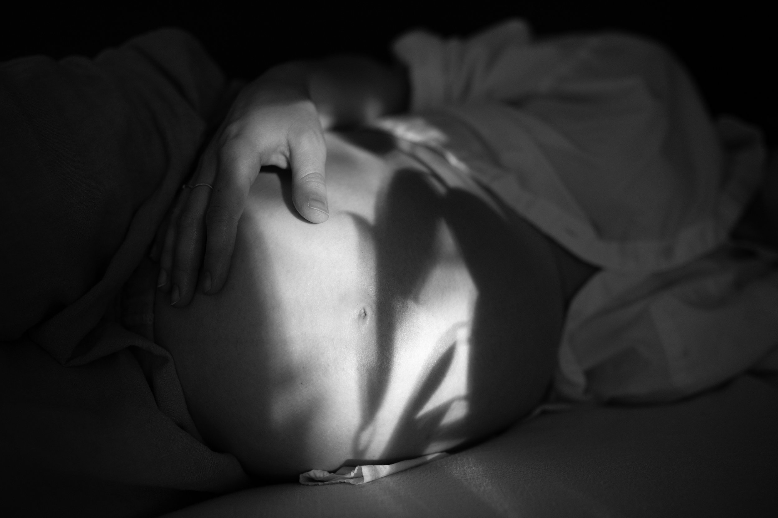 Black and white photograph of a person lying down with their hand resting on their stomach, showing a side profile of their face and torso, partially covered by a blanket or clothing.