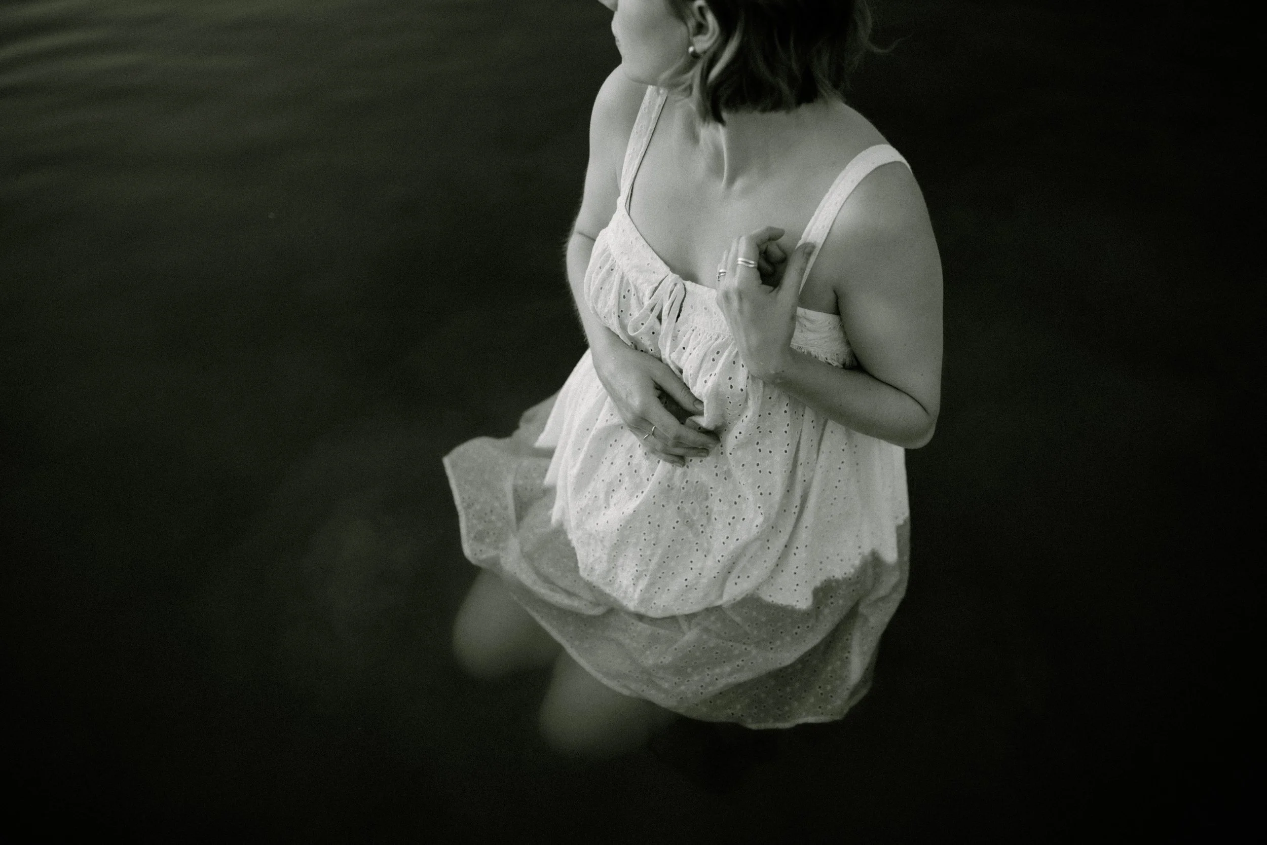 A pregnant woman in a light-colored dress is sitting on a boat or dock, with water visible in the background, seen from an overhead angle.