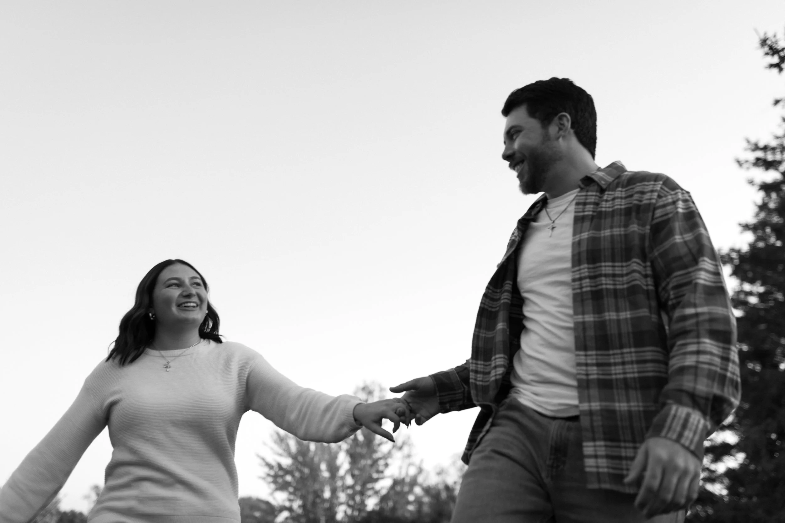 Two people, a woman and a man, holding hands and smiling at each other outdoors in black-and-white, with trees in the background.