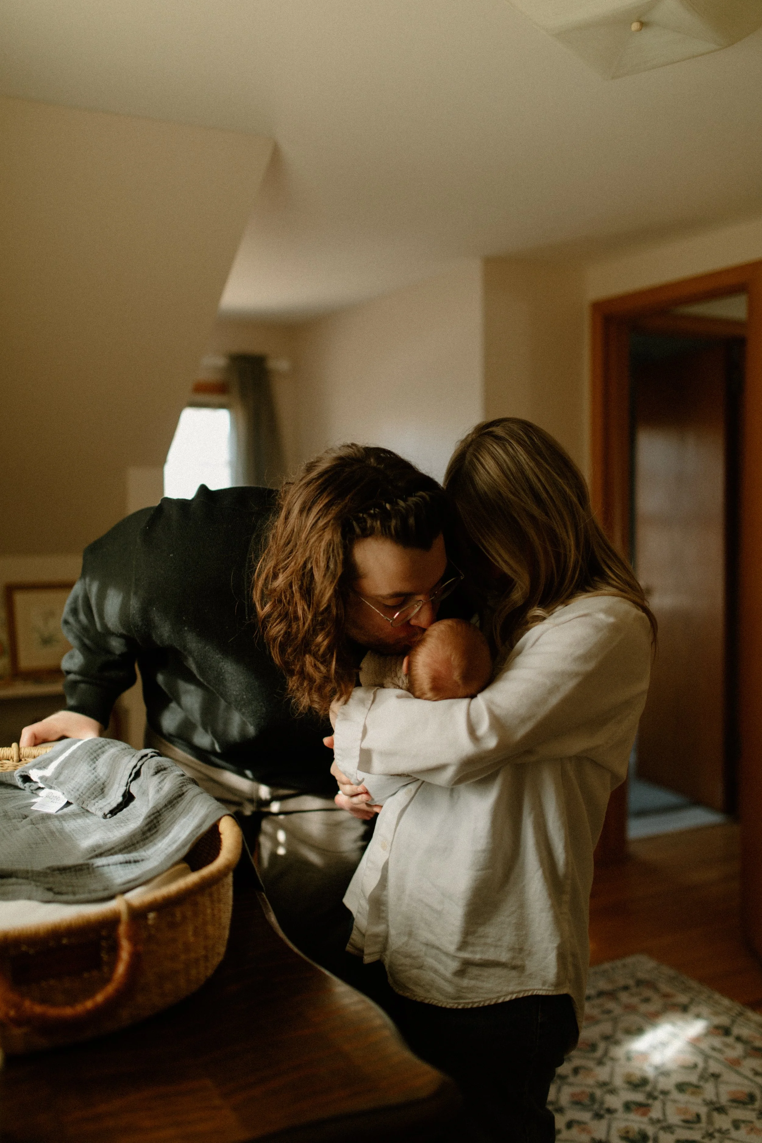 A young could and a baby in a cozy home setting, with the young mother holding the baby close while the father leans in for a kiss on the baby's head.