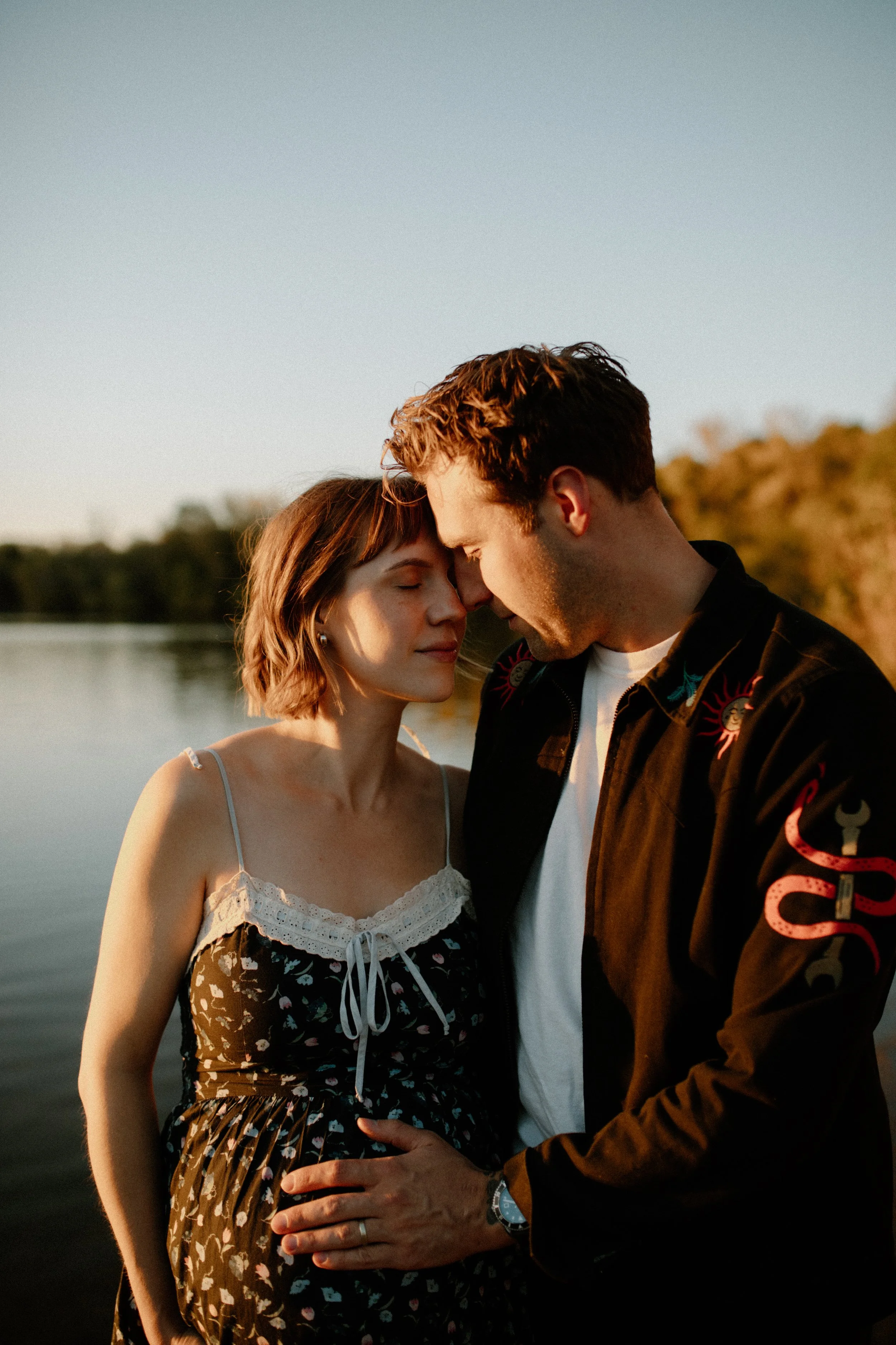 A young couple standing close together by a body of water during sunset, with the man touching the woman's belly and both with their foreheads touching, sharing an intimate moment.