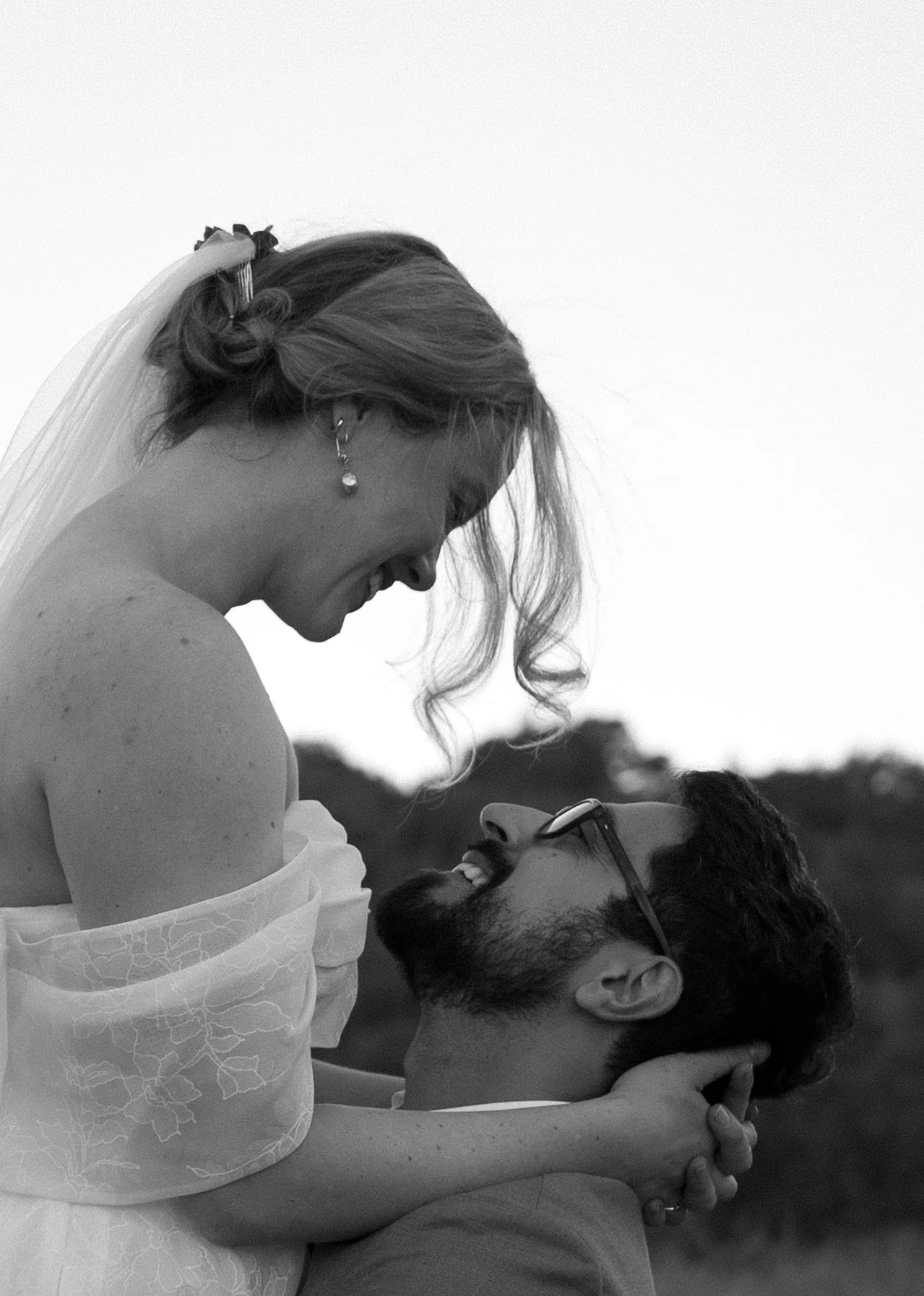 A black and white photo of a smiling couple, with the woman sitting on the man's shoulders and leaning forward to look at him, while he holds her and looks up at her. The woman has wavy hair and is wearing earrings, and the man has glasses and a bear