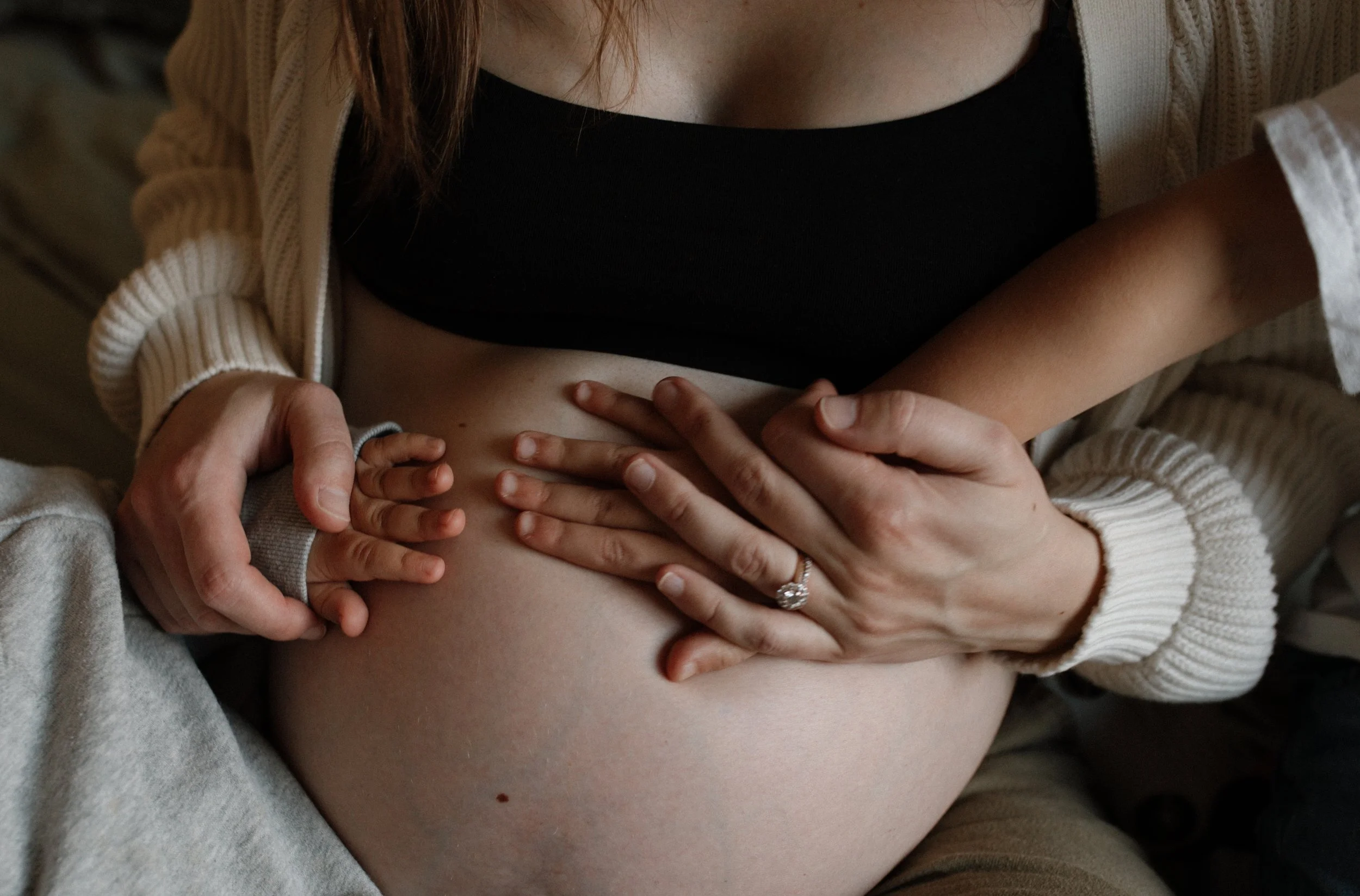 Close-up of a woman’s pregnant belly with several hands touching her stomach, one of which is wearing a ring.