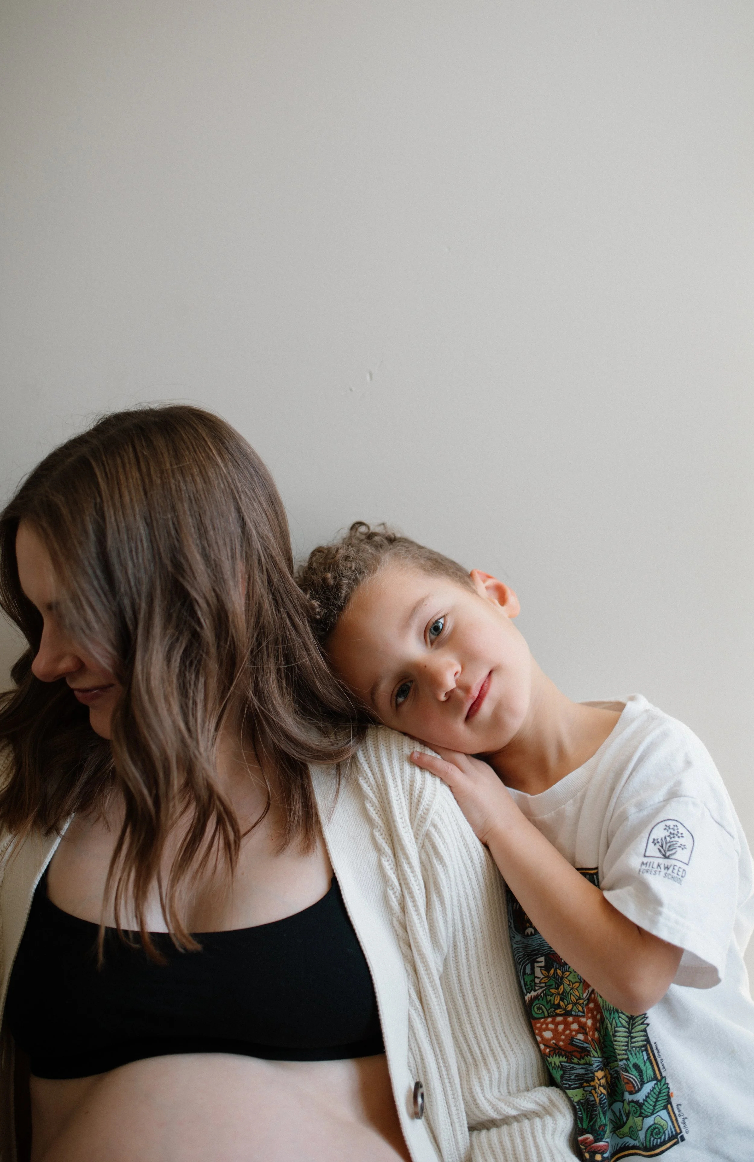A woman with long brown hair and a black tank top underneath a white cardigan is holding a young boy with curly hair and a colorful shirt. The boy is resting his head on her shoulder and looking at the camera. Both are against a plain light gray wall