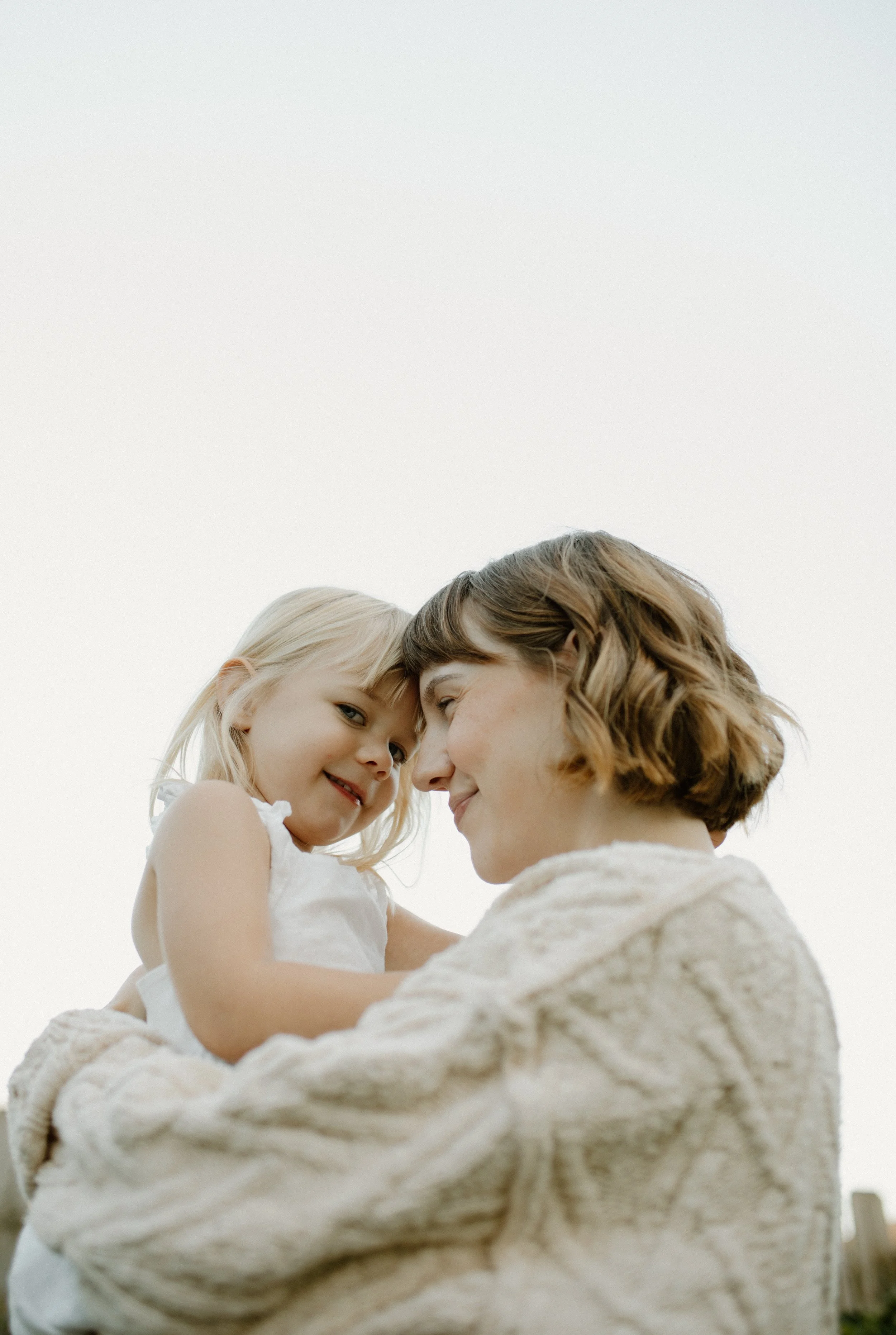 A woman holding a young girl close with their faces touching, both smiling softly, against a plain light sky background.