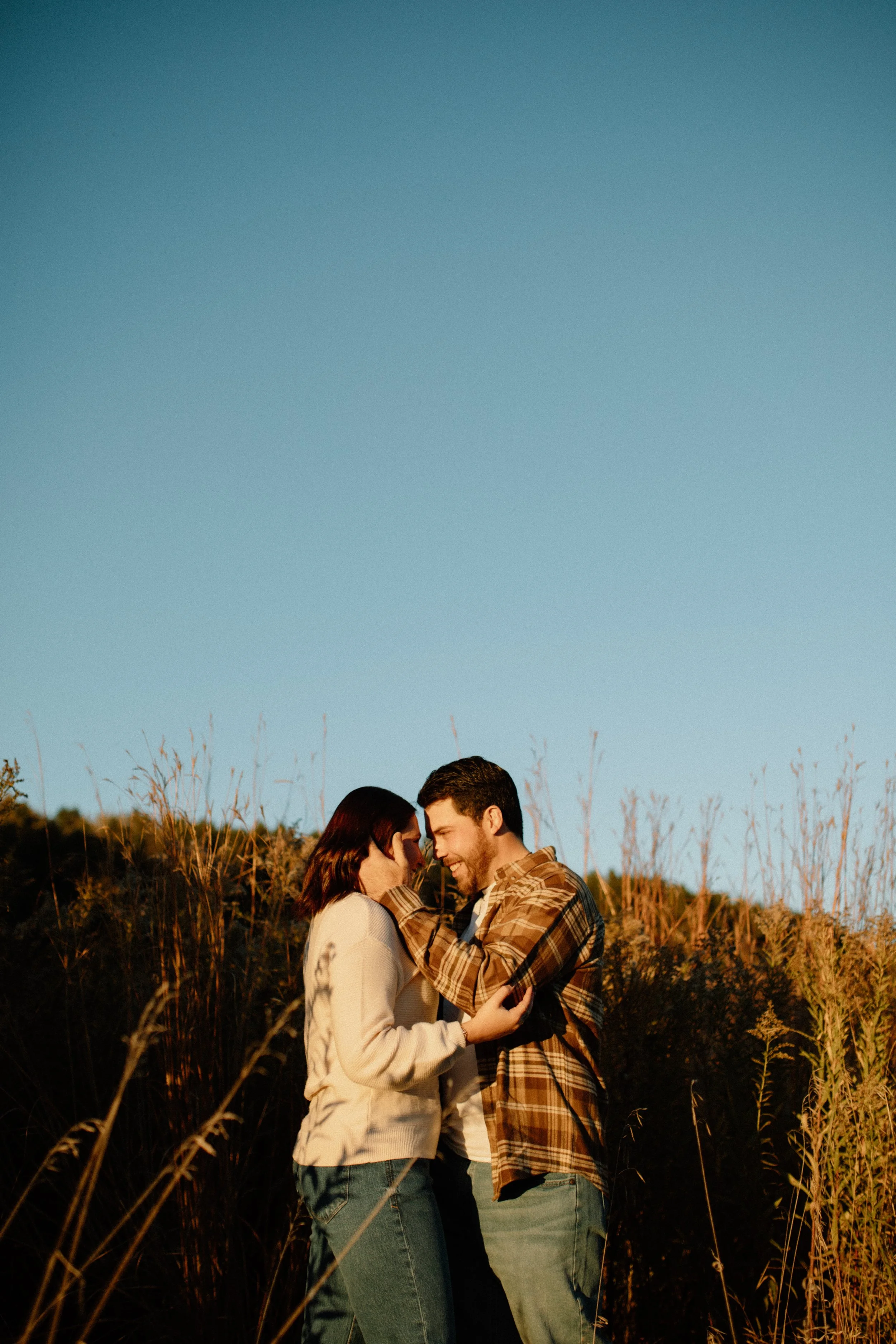 A young couple standing close together in a field at sunset, touching foreheads and smiling at each other, surrounded by tall grass and plants.