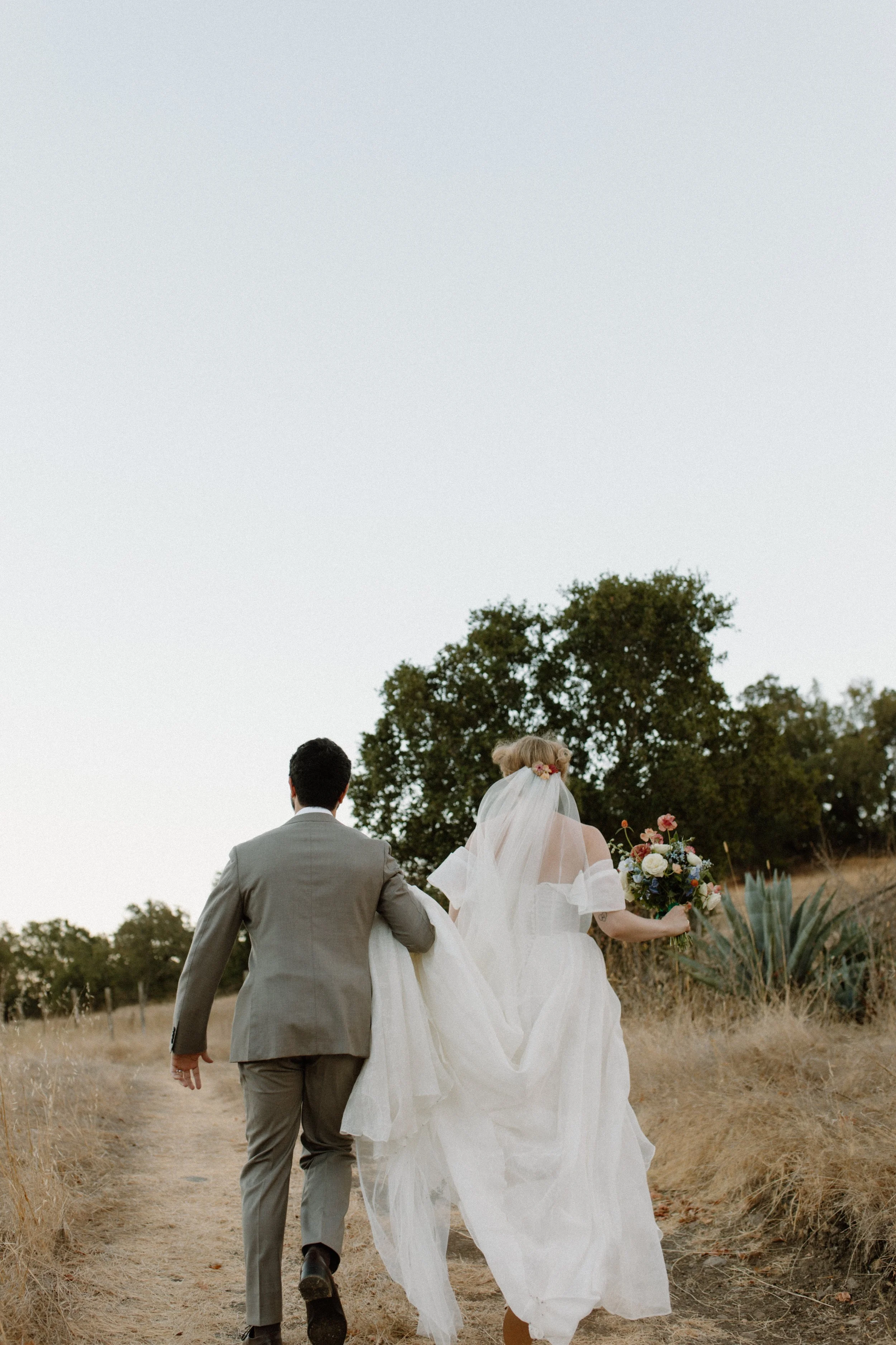 A bride and groom walking on a dirt path outdoors, with trees and grass, during daytime. The bride is holding a bouquet of flowers and wearing a white wedding dress with a veil, and the groom is in a gray suit. They are walking away from the camera.